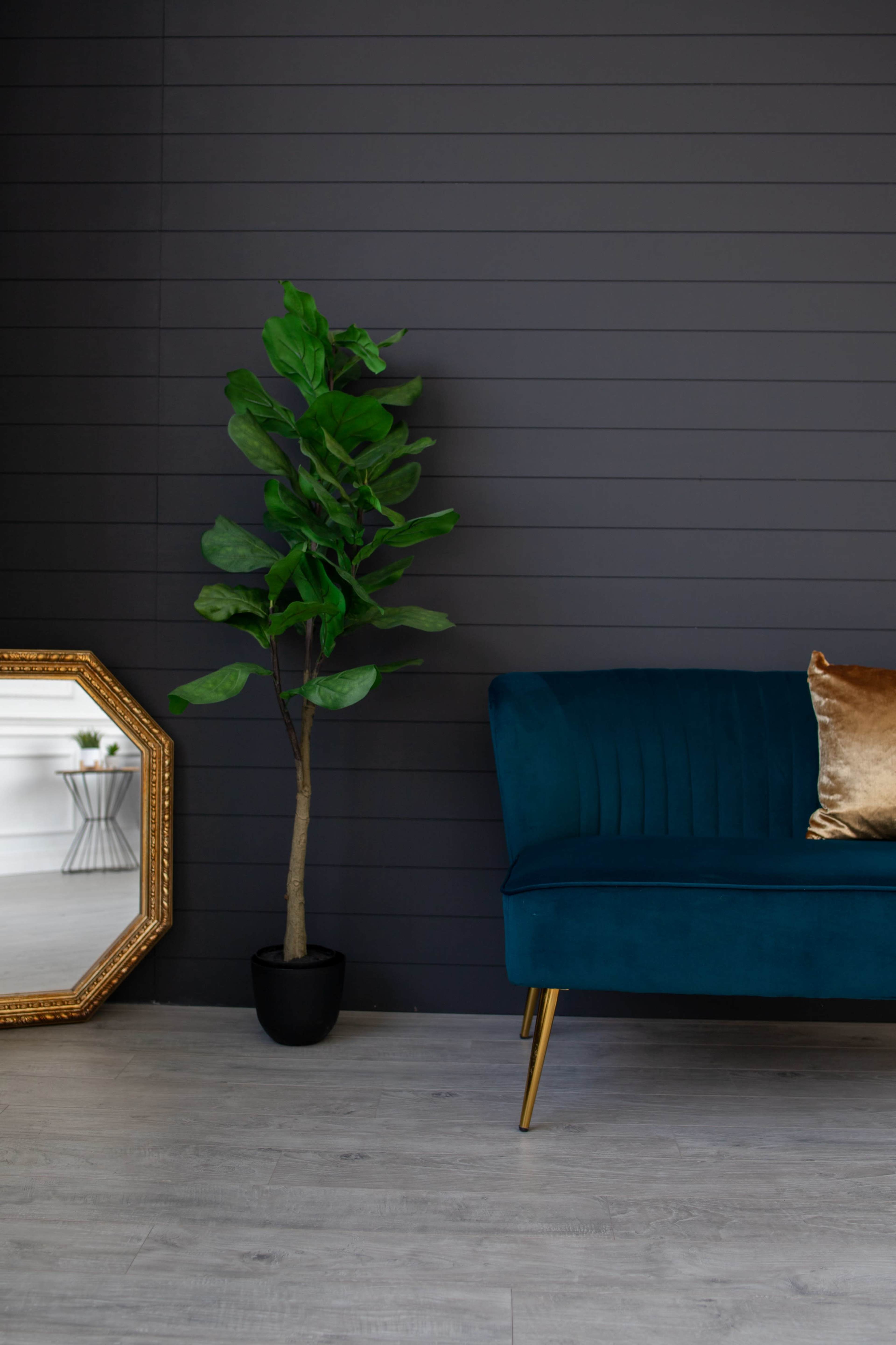 A teal sofa with a gold pillow is positioned next to a potted plant and a decorative mirror against a dark paneled wall.