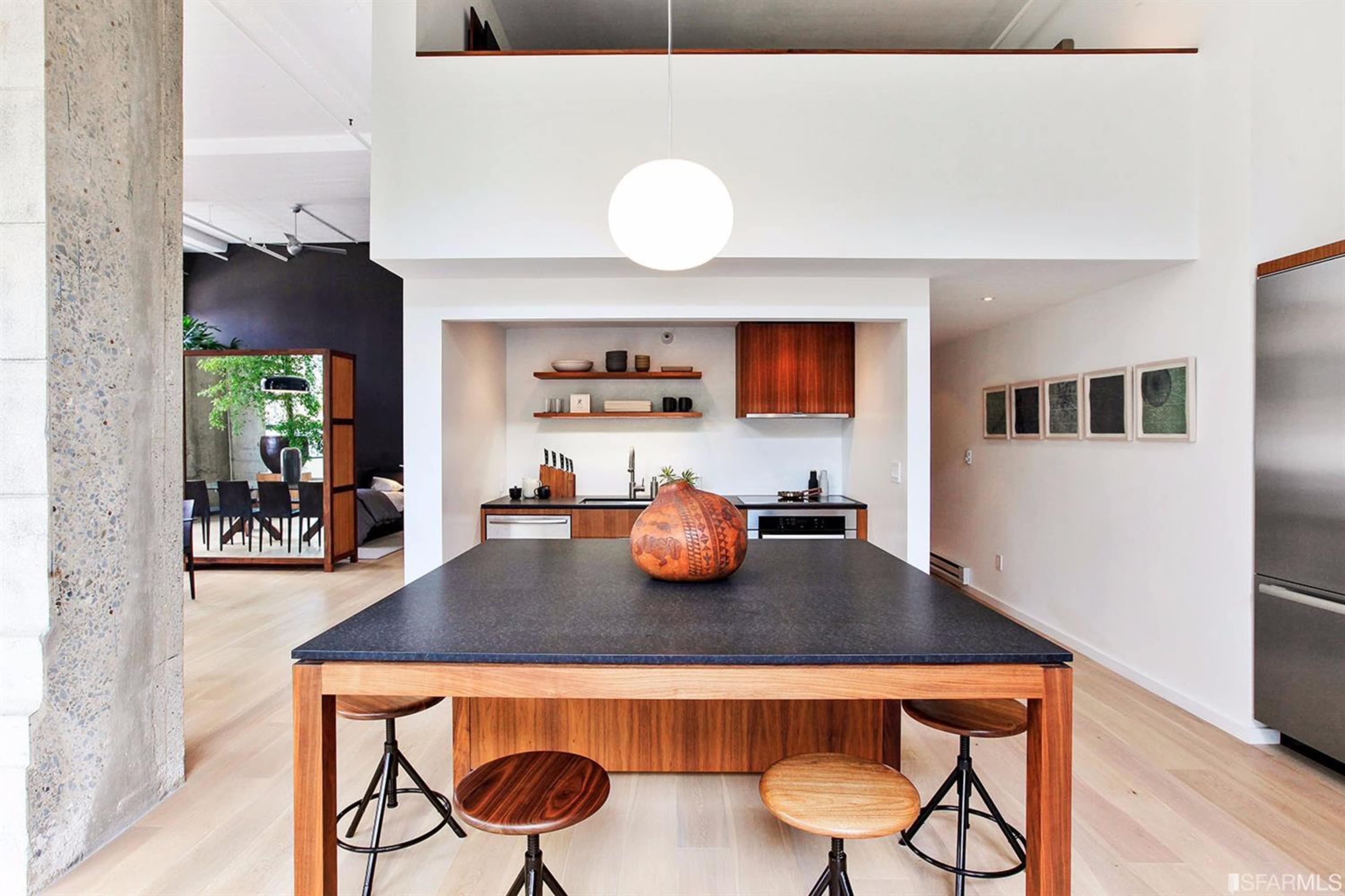 A modern kitchen features a dark countertop, wooden cabinetry, and a round pendant light above a dining table with stools.