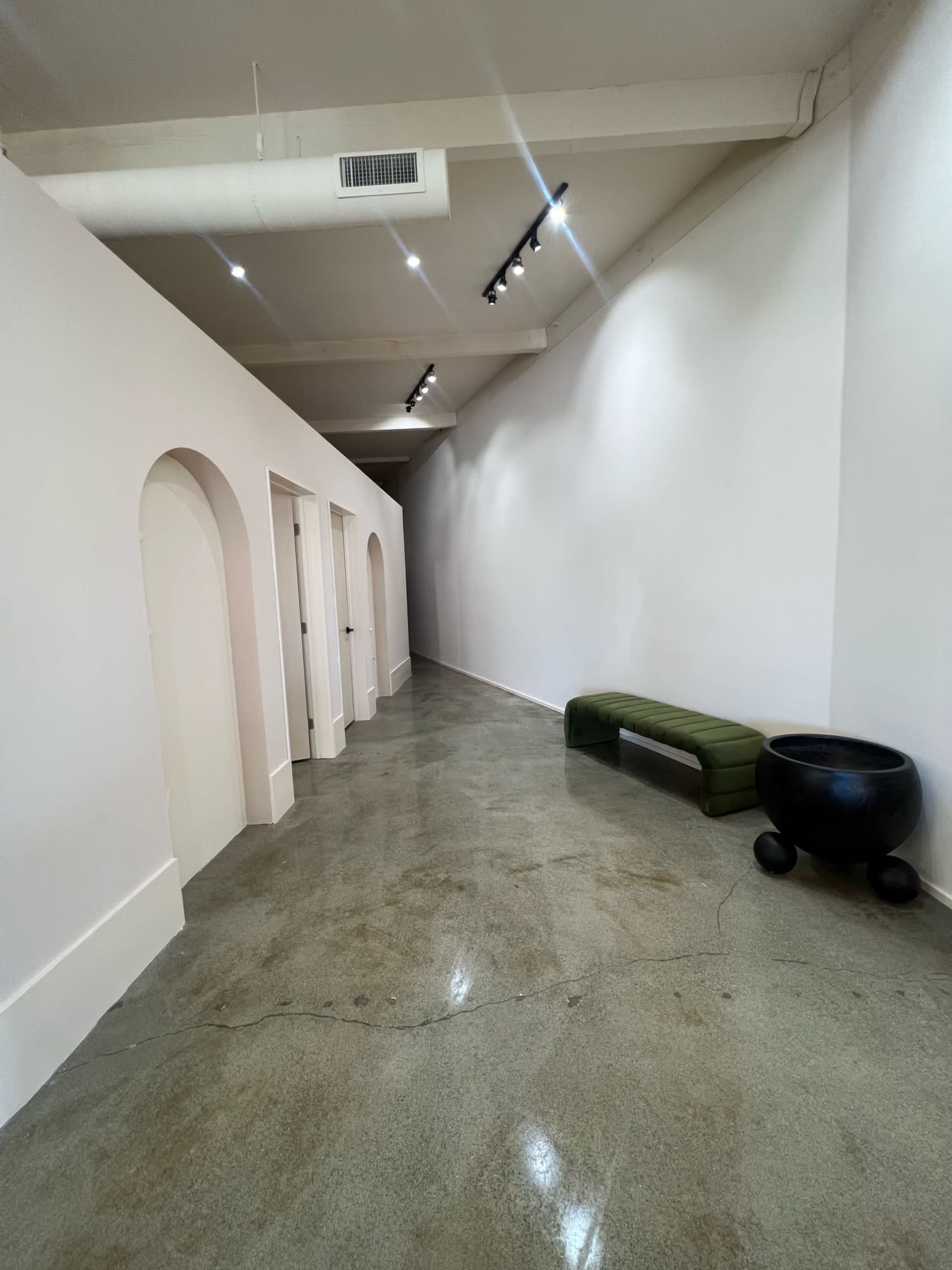 A narrow hallway features a smooth concrete floor, archway doorways, and minimalistic seating along the wall.