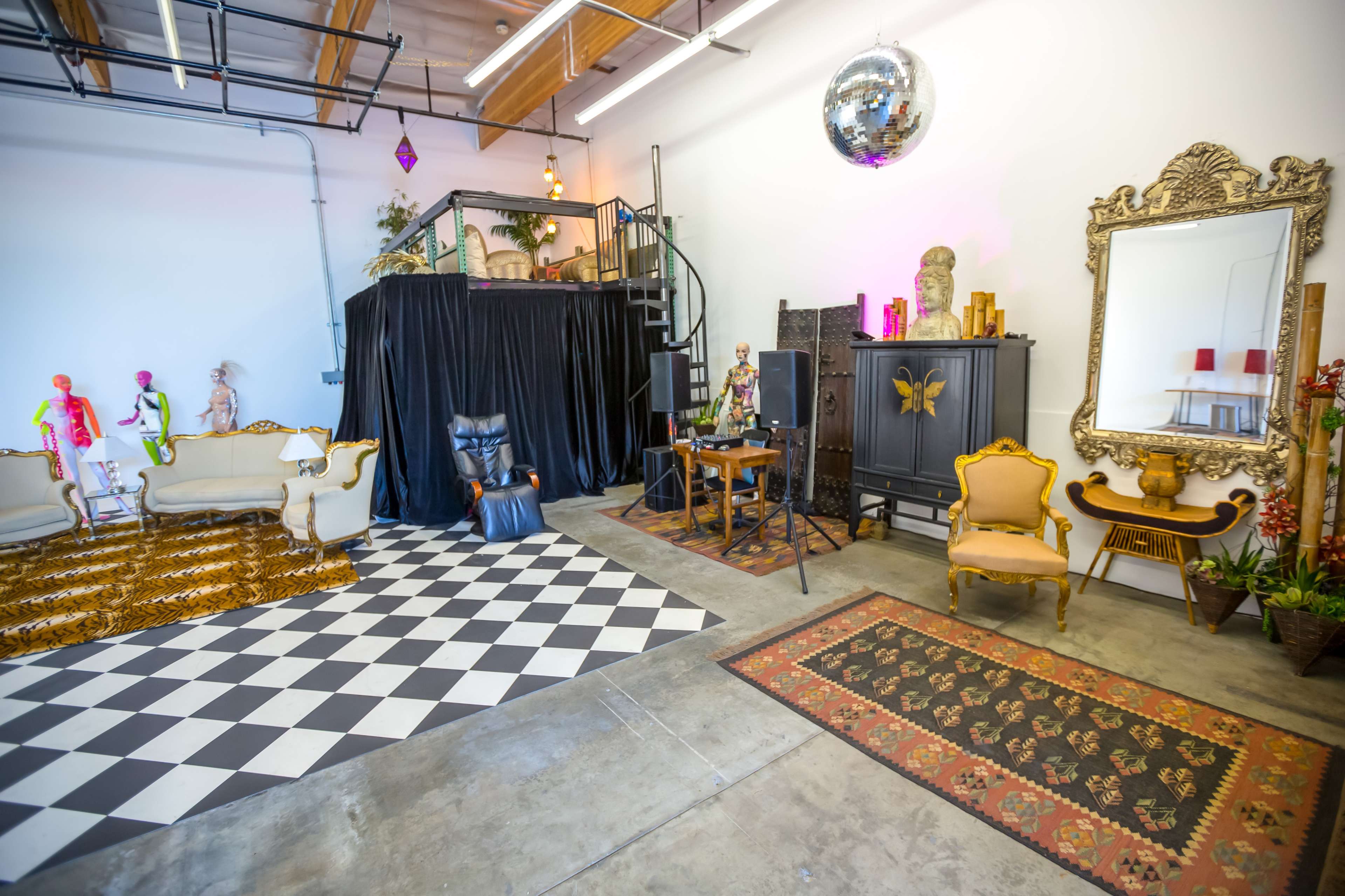 The image shows a spacious art studio featuring a black and white checkered floor, various vintage furniture pieces, a mirrored wall, and a staircase leading to a raised platform.