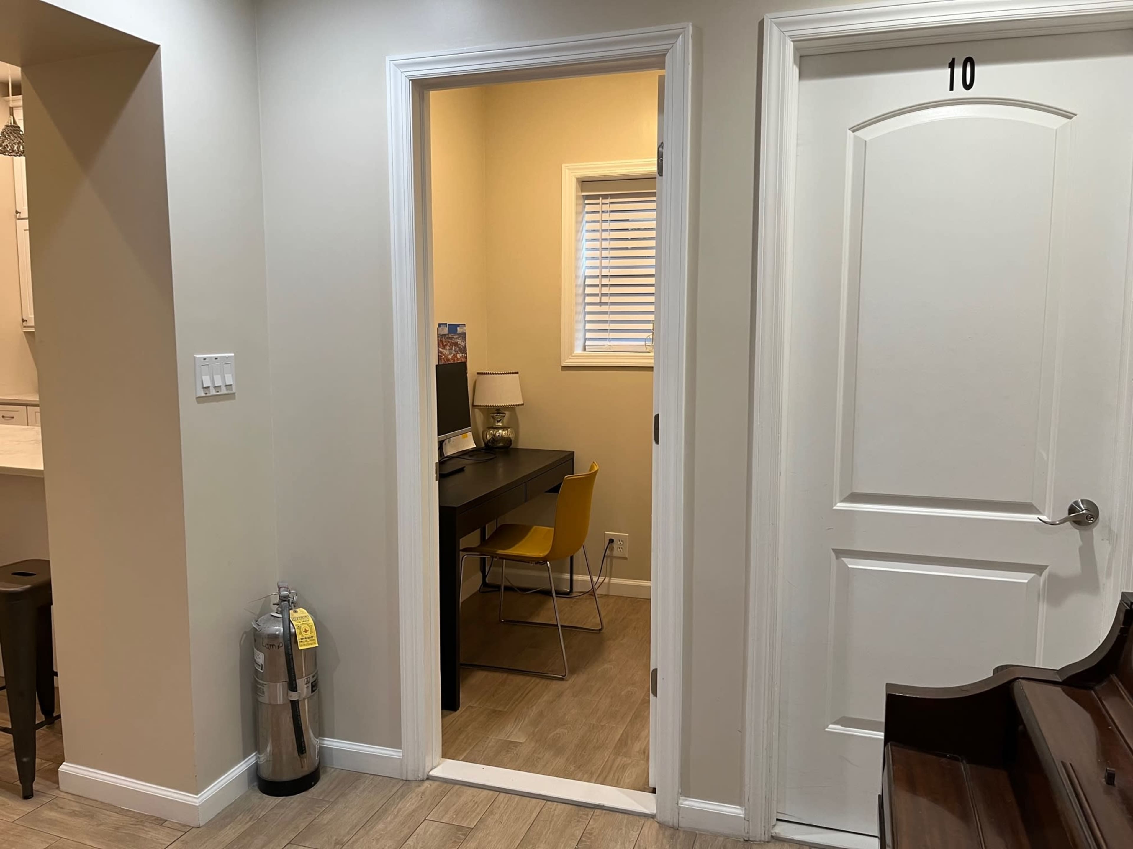 A narrow hallway leading to a small office space with a desk and a chair, next to a closed door labeled "10" and a fire extinguisher mounted on the wall.