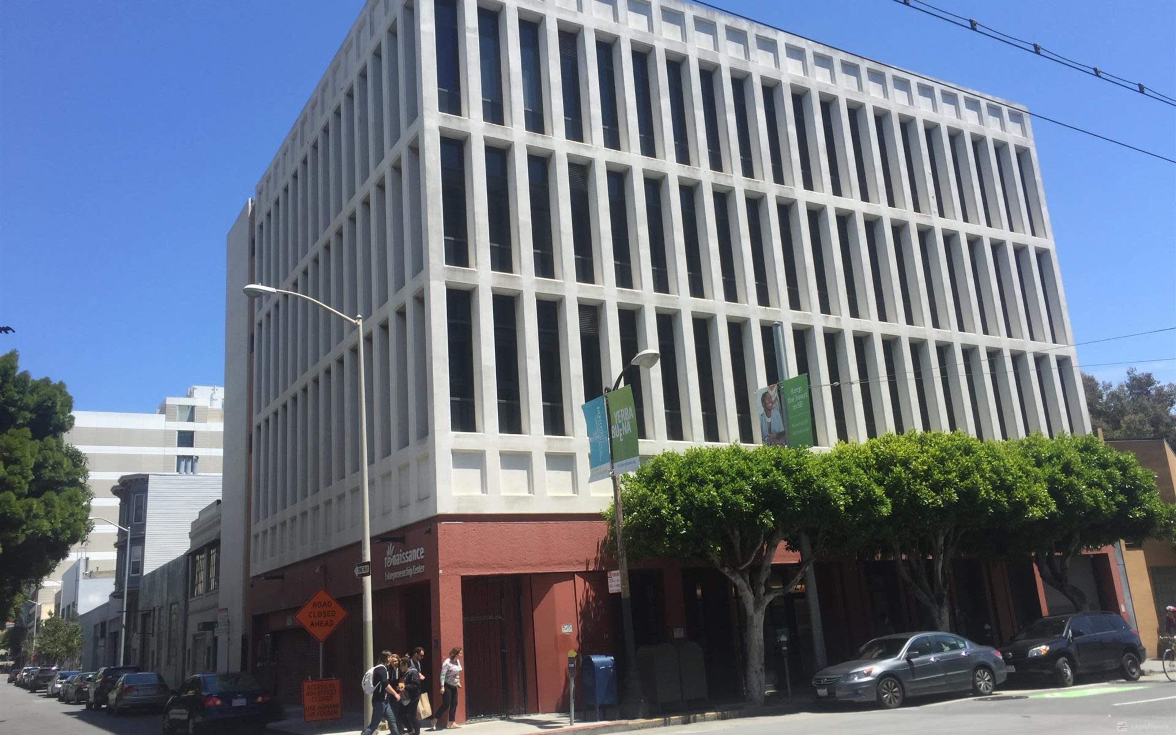 A five-story building with a modern façade stands at the corner of a bustling street, flanked by parked cars and pedestrians.