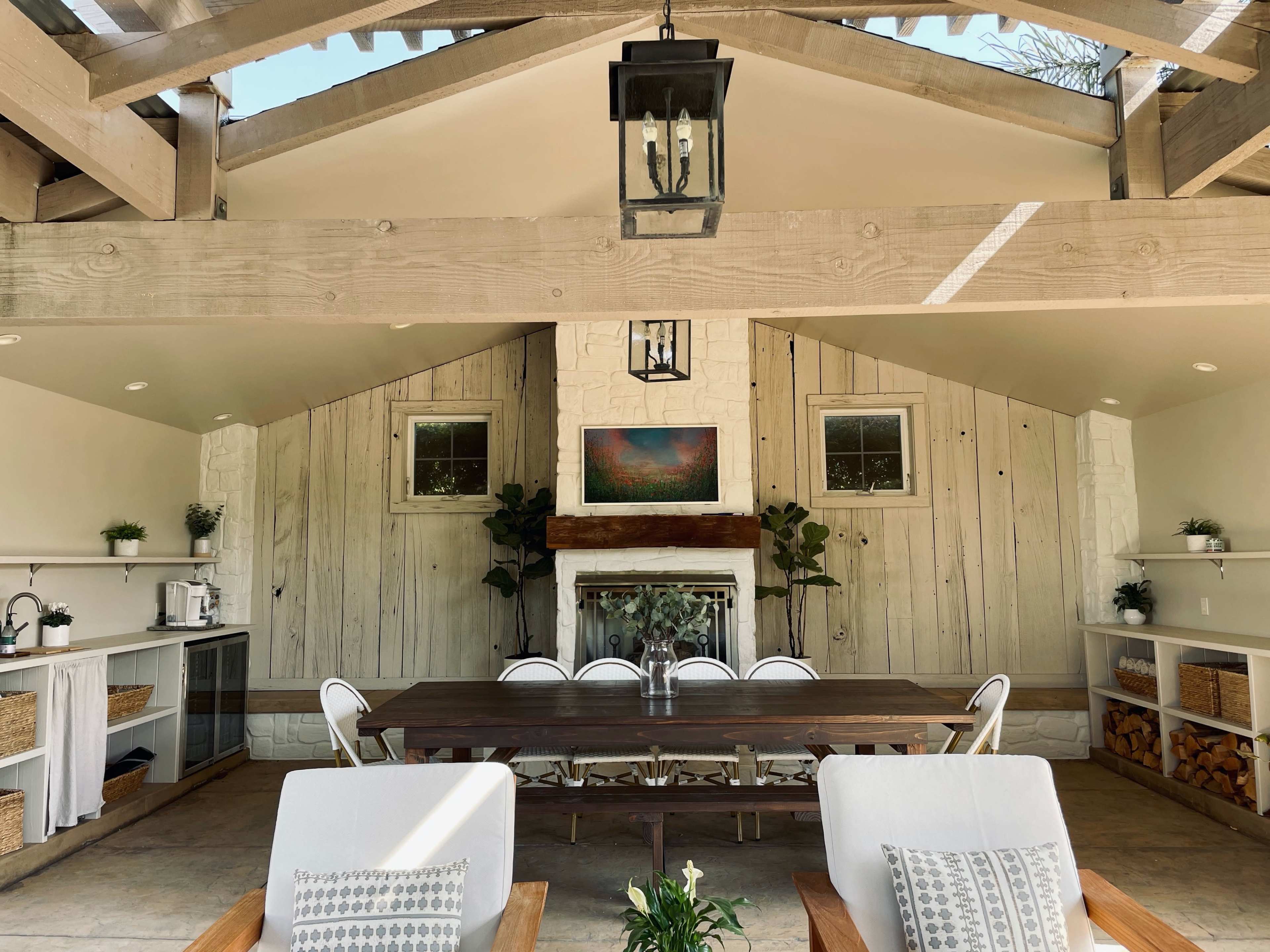 The image shows a spacious, well-lit indoor seating area with a long wooden dining table, surrounded by chairs and decorated with potted plants and artwork on the walls.