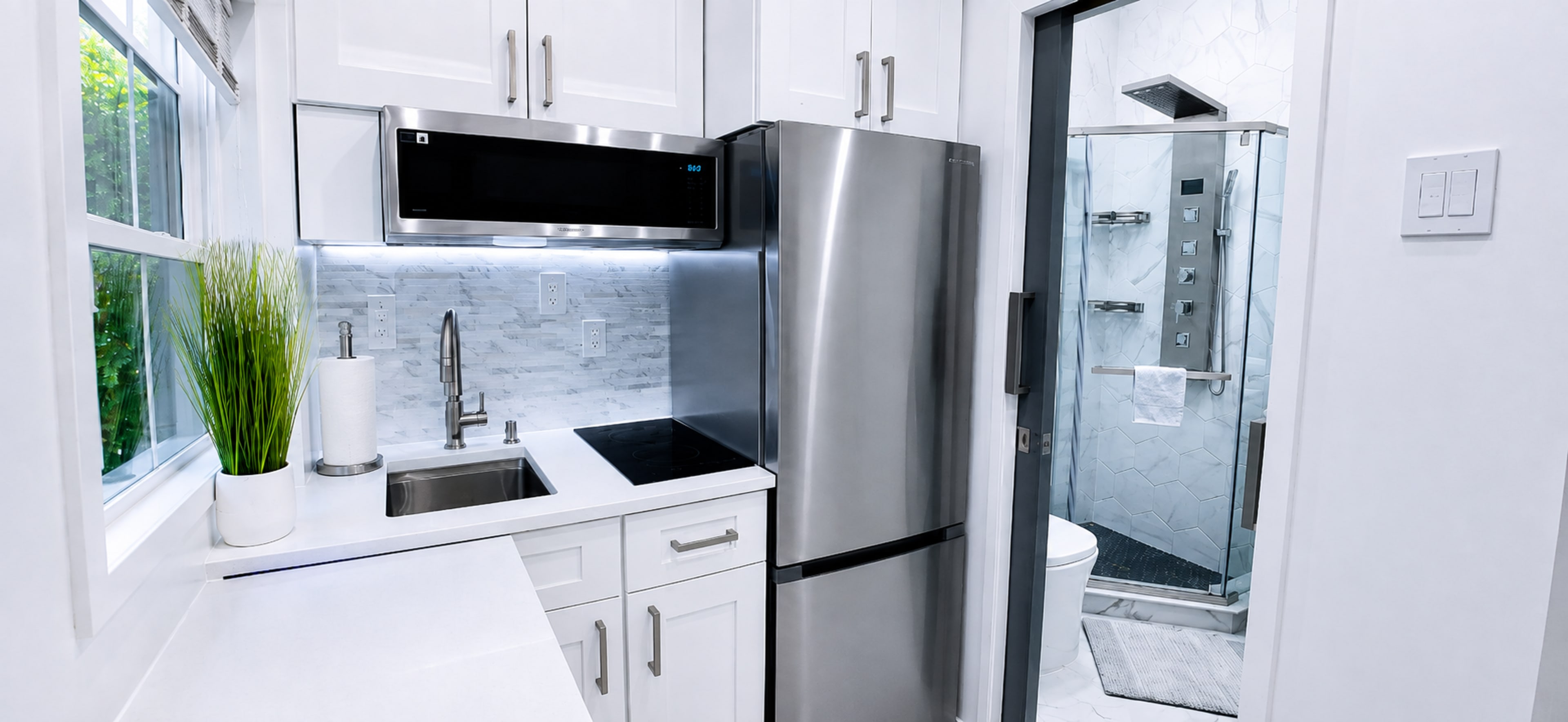 The image shows a modern kitchen with a stainless steel refrigerator, a sink, and a small cooking area adjacent to a glass-enclosed bathroom.