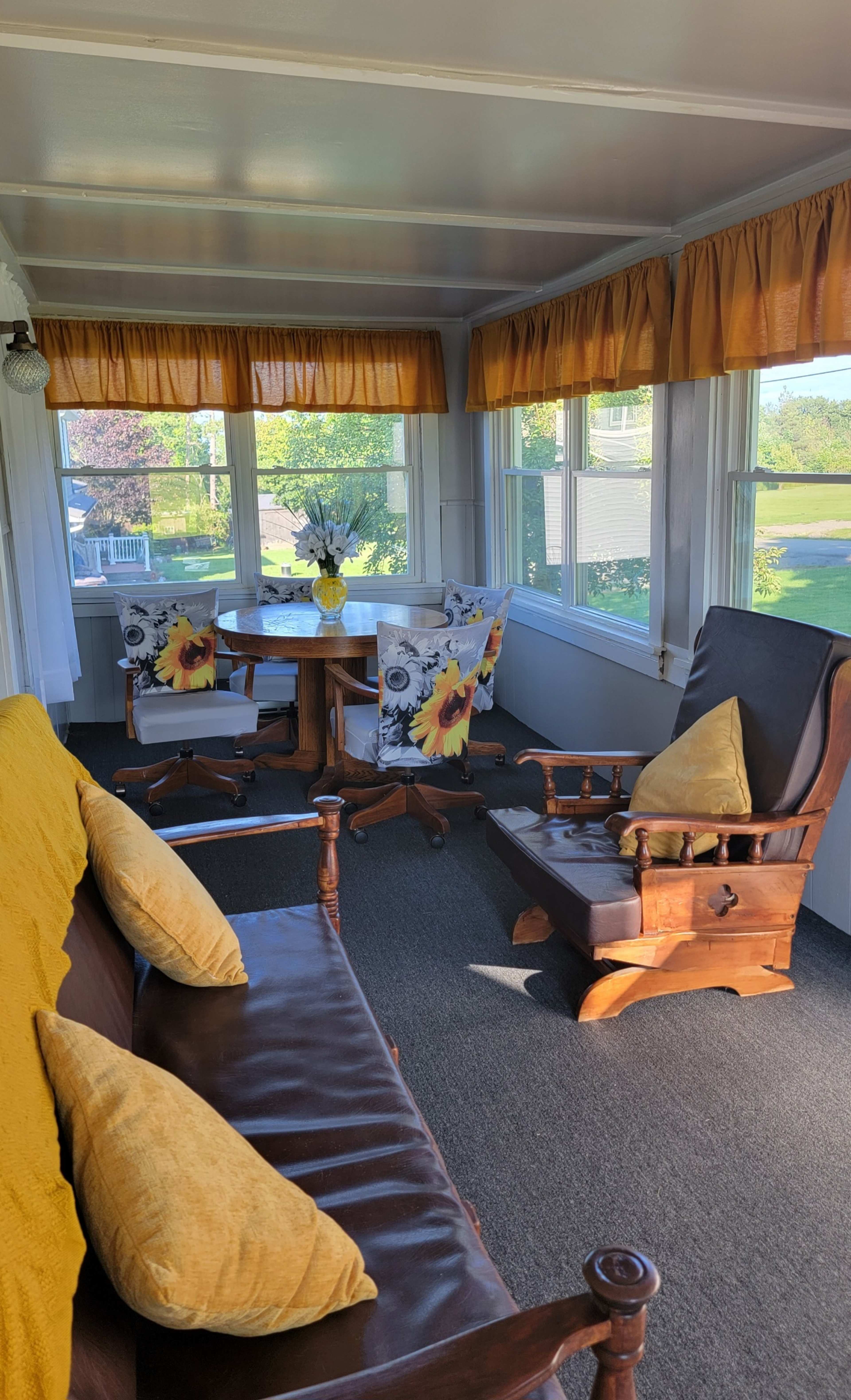 A sunlit enclosed porch features a brown sofa with yellow pillows, a wooden table, and chairs adorned with floral upholstery.