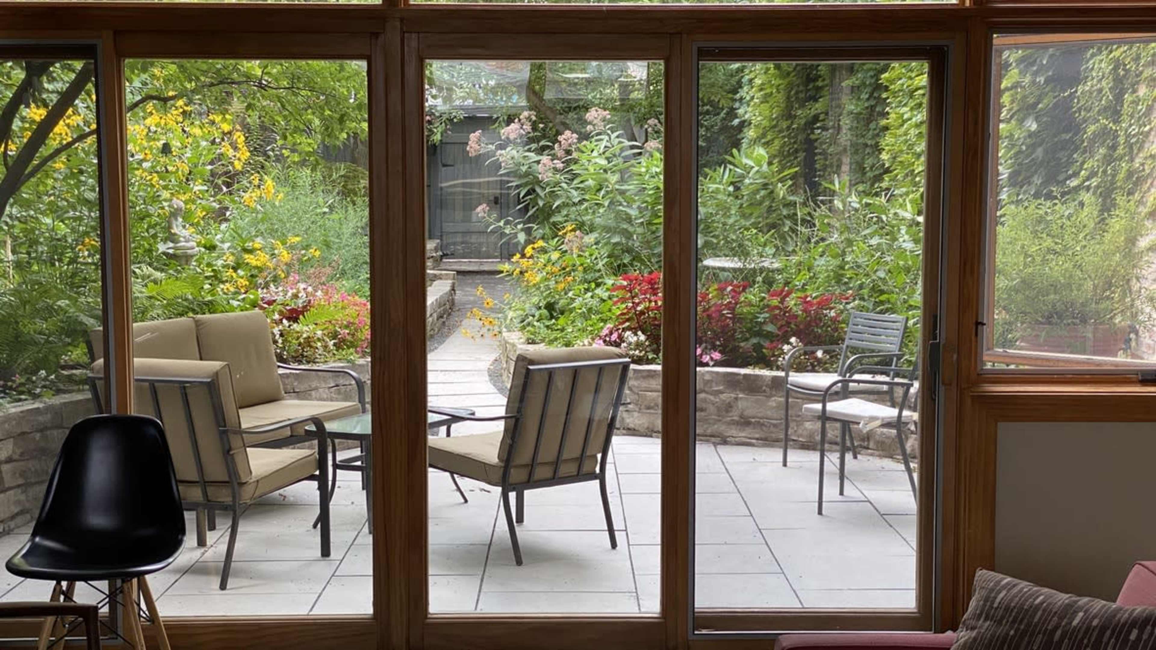 A patio area with four chairs and a small table is visible through large glass windows, surrounded by a garden filled with flowers and greenery.