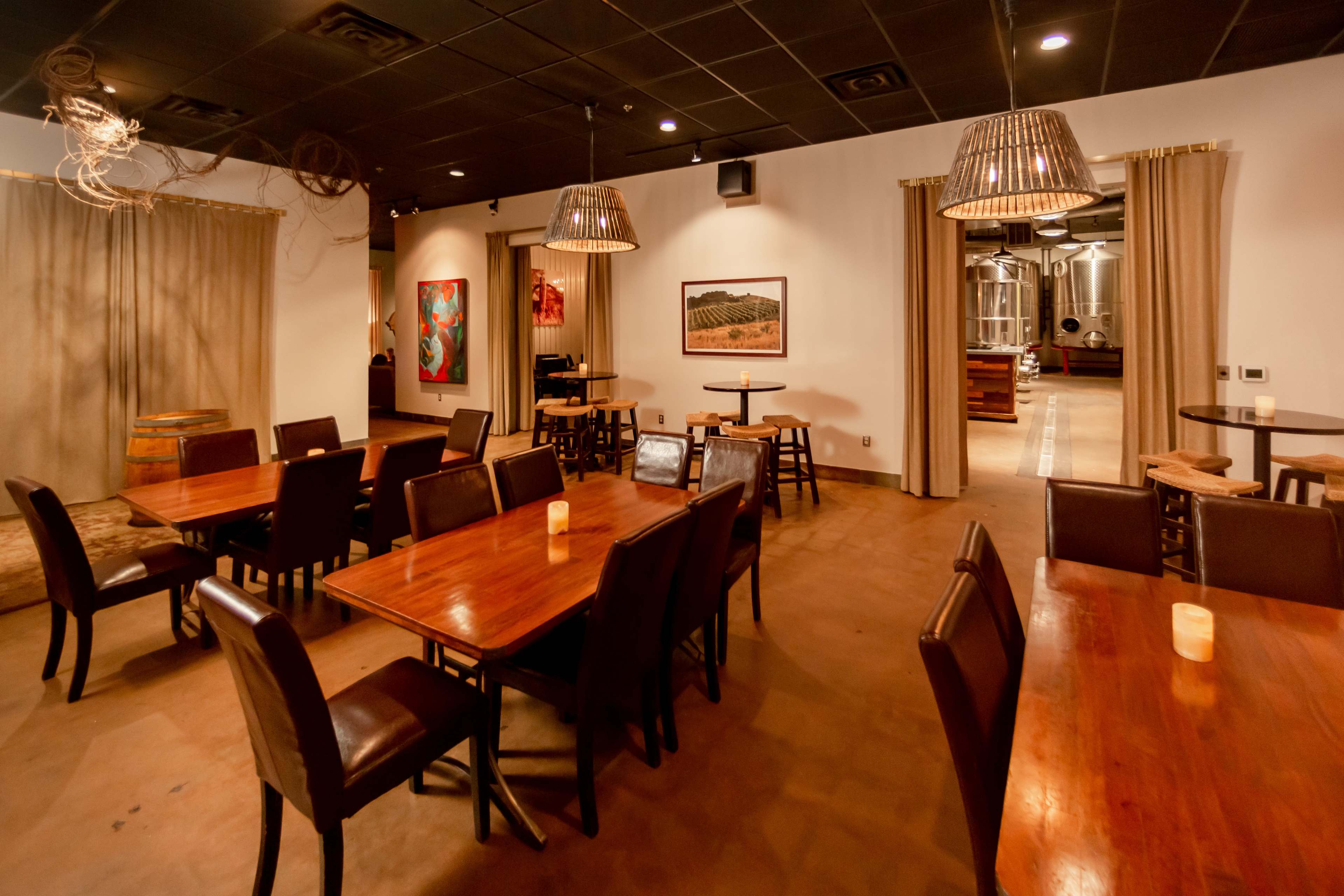 A spacious dining area with wooden tables and chairs, featuring dim lighting from hanging pendant lamps, and a view of brewing equipment in the adjacent room.
