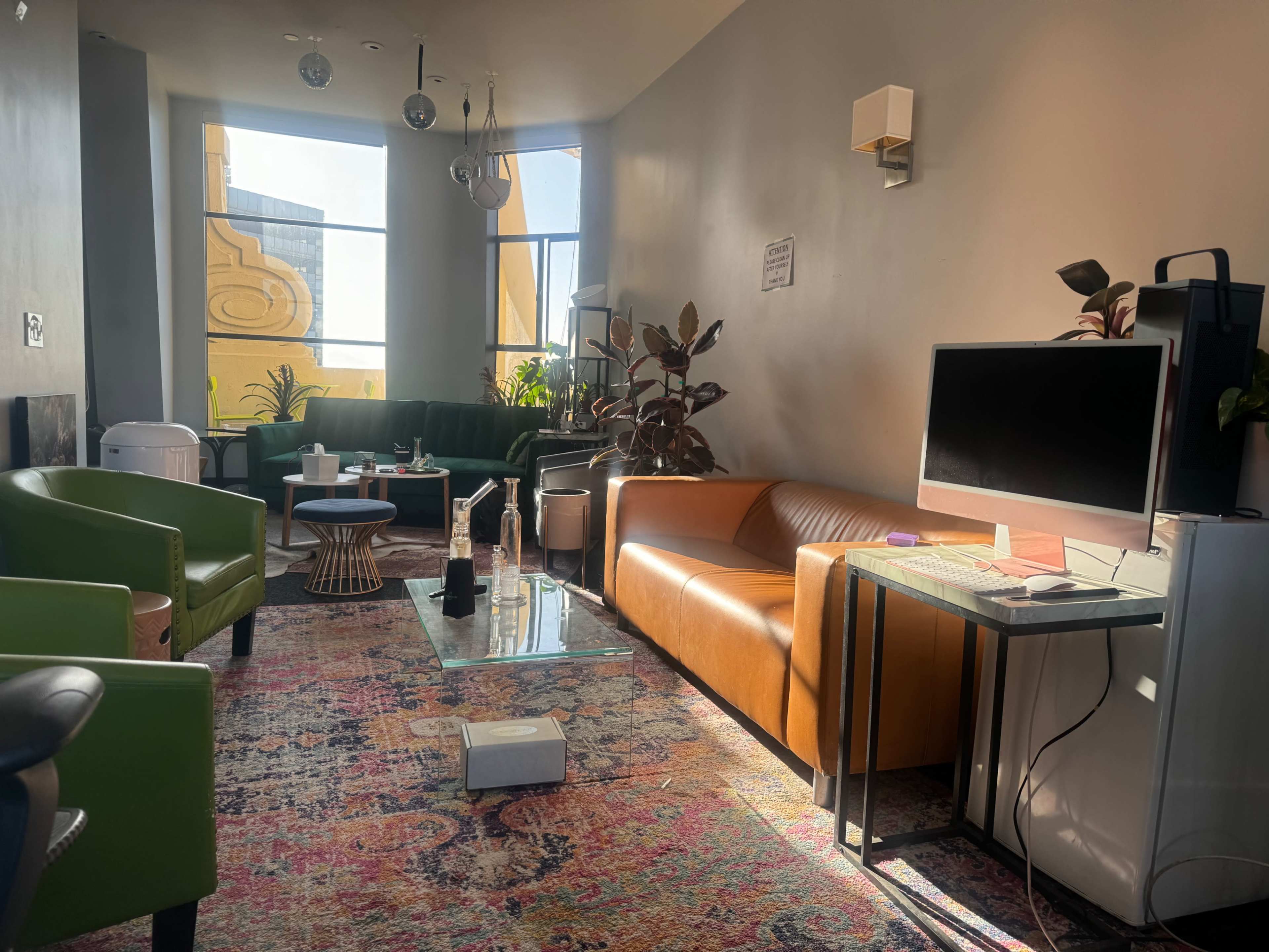 A modern lounge area with a brown leather sofa, green armchairs, a glass coffee table, and large windows letting in natural light.