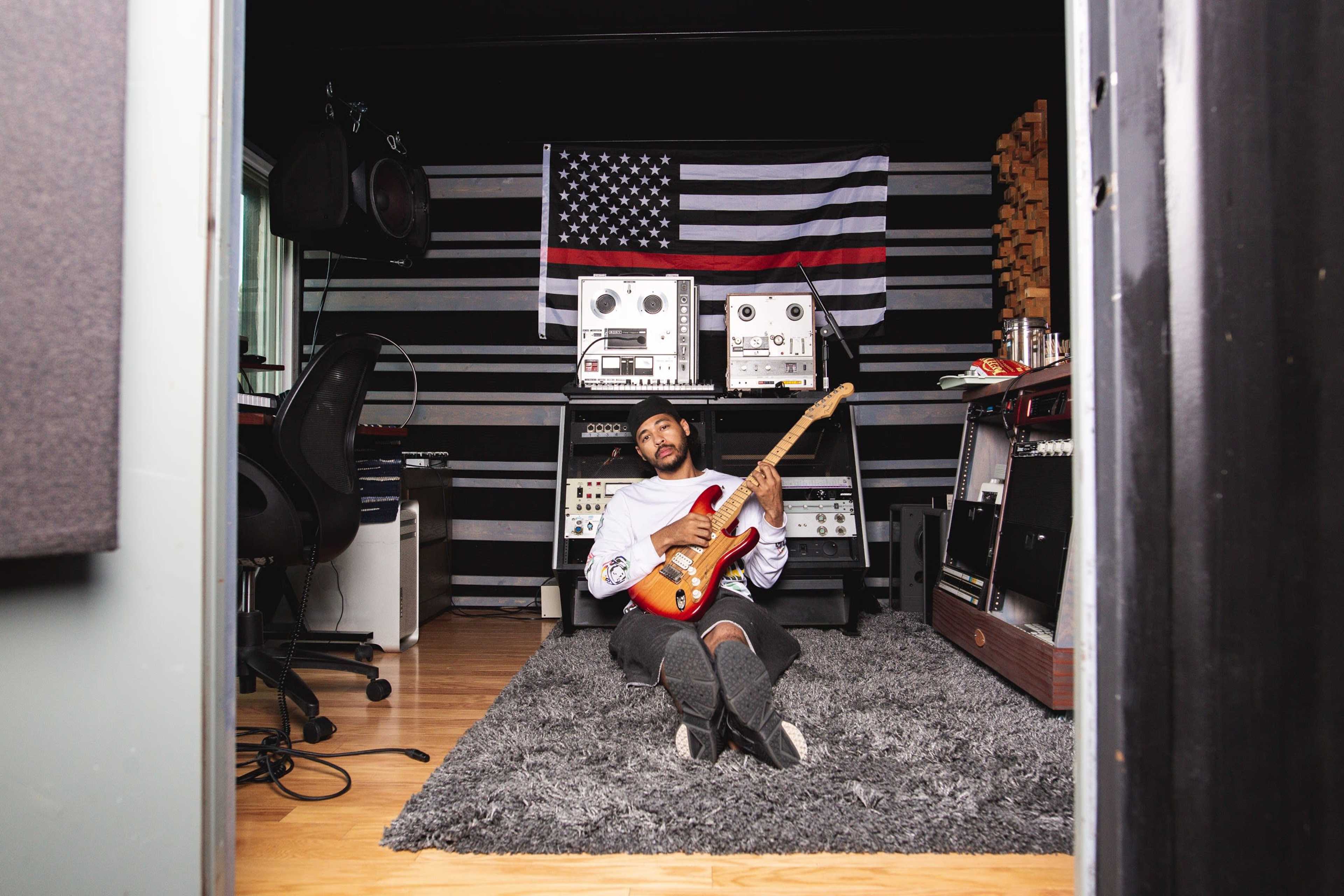 A musician sits on the floor of a recording studio, playing an electric guitar in front of vintage audio equipment and an American flag.