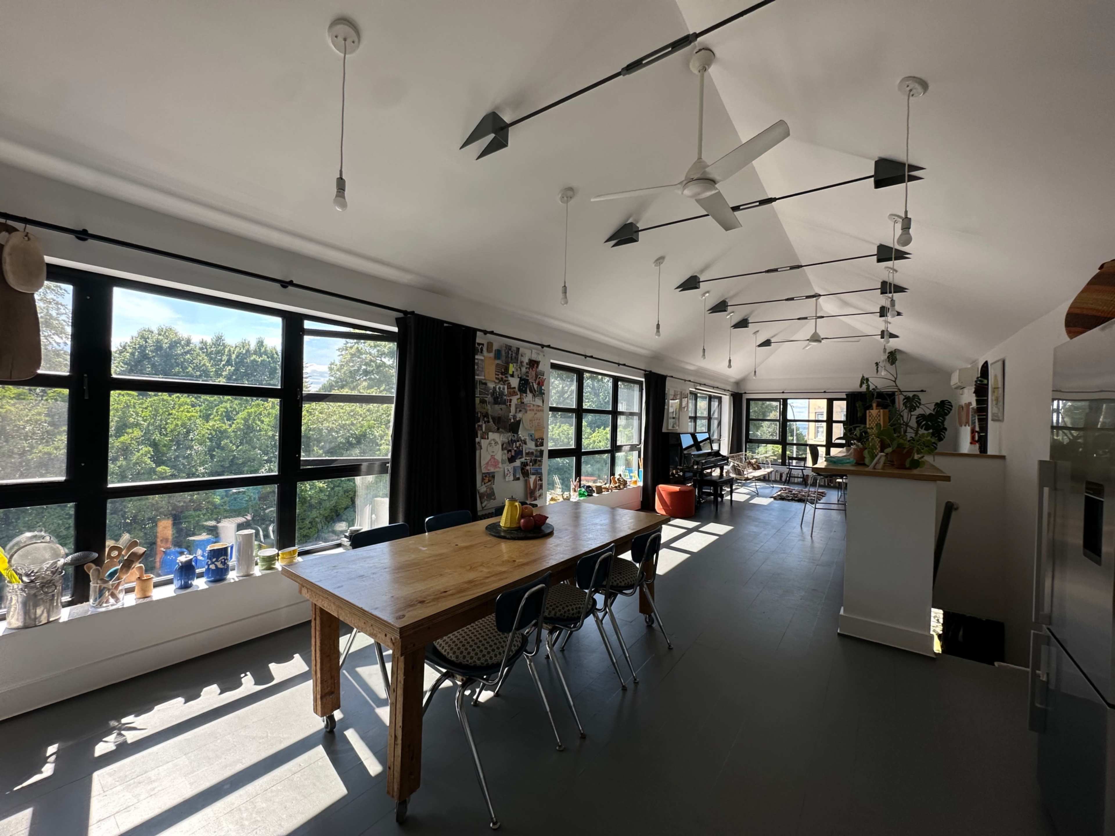 The image shows a spacious, modern dining area with large windows, a wooden table surrounded by chairs, and overhead fans, leading to a living space filled with natural light.