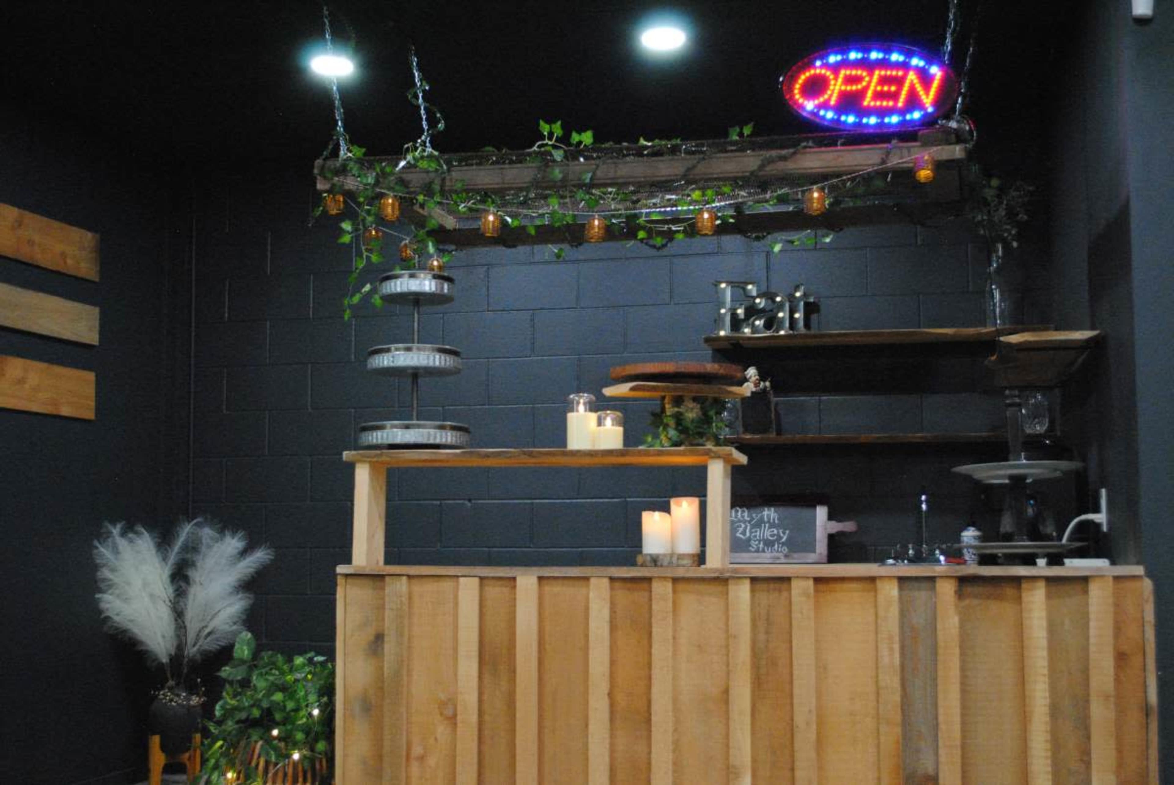 The image shows a rustic wooden reception desk with an illuminated "OPEN" sign overhead, surrounded by decorative plants and shelves.