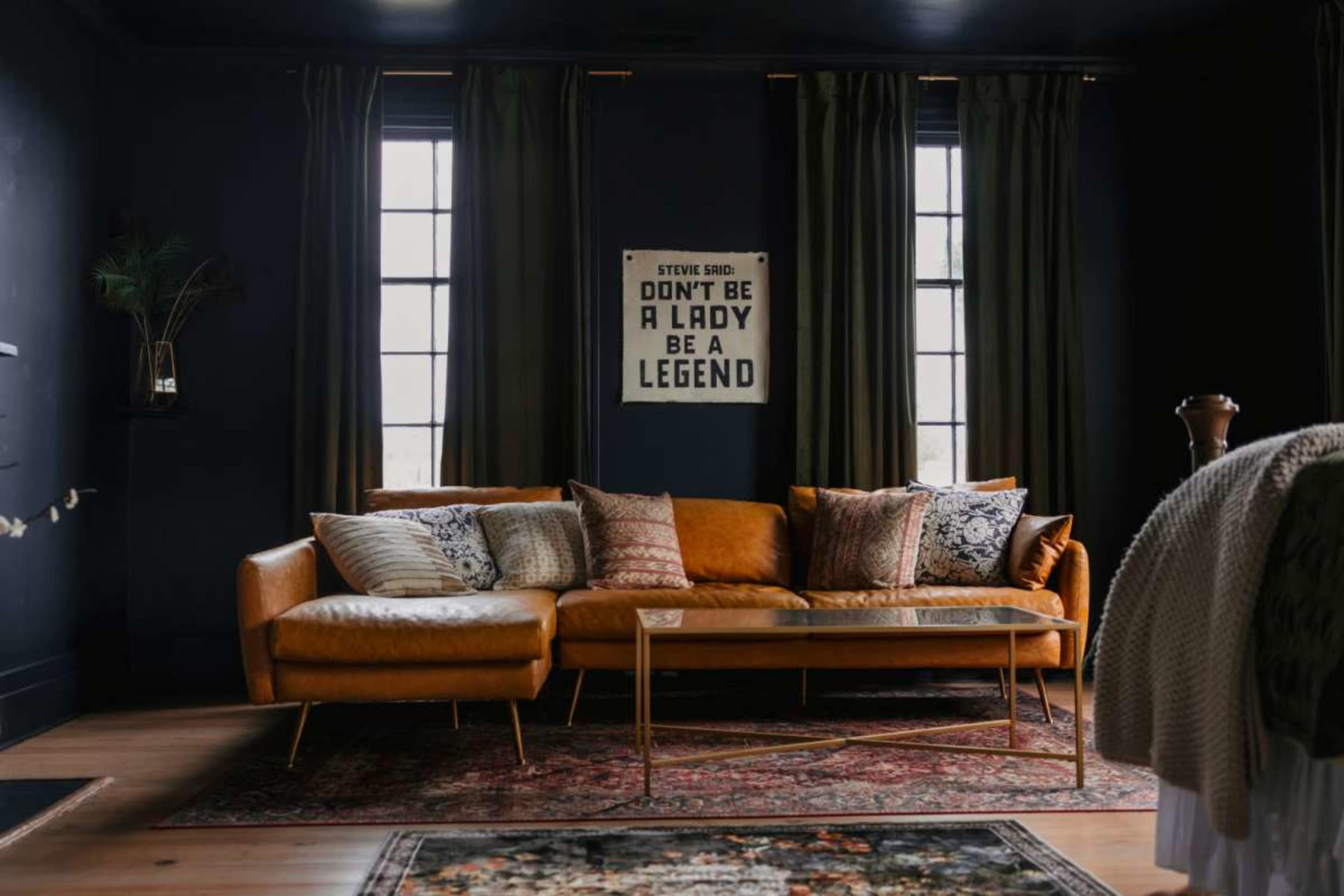 A modern living room features a mustard-colored sectional sofa adorned with decorative pillows, a glass coffee table, and a wall hanging with an inspirational quote, set against dark walls and large windows draped with green curtains.