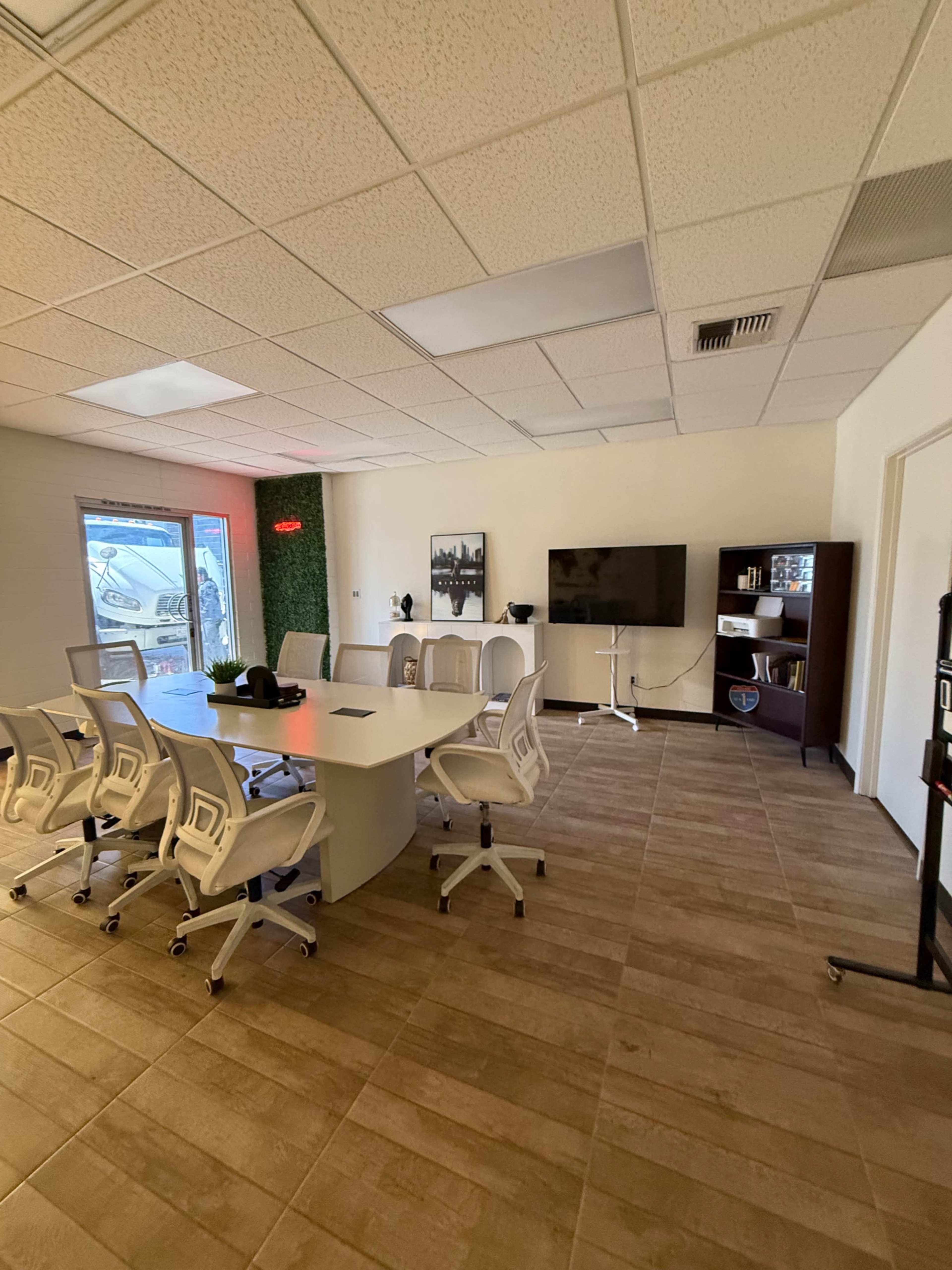 The image shows a modern conference room with a large oval table surrounded by white ergonomic chairs, a television on the wall, and a bookshelf in the corner.