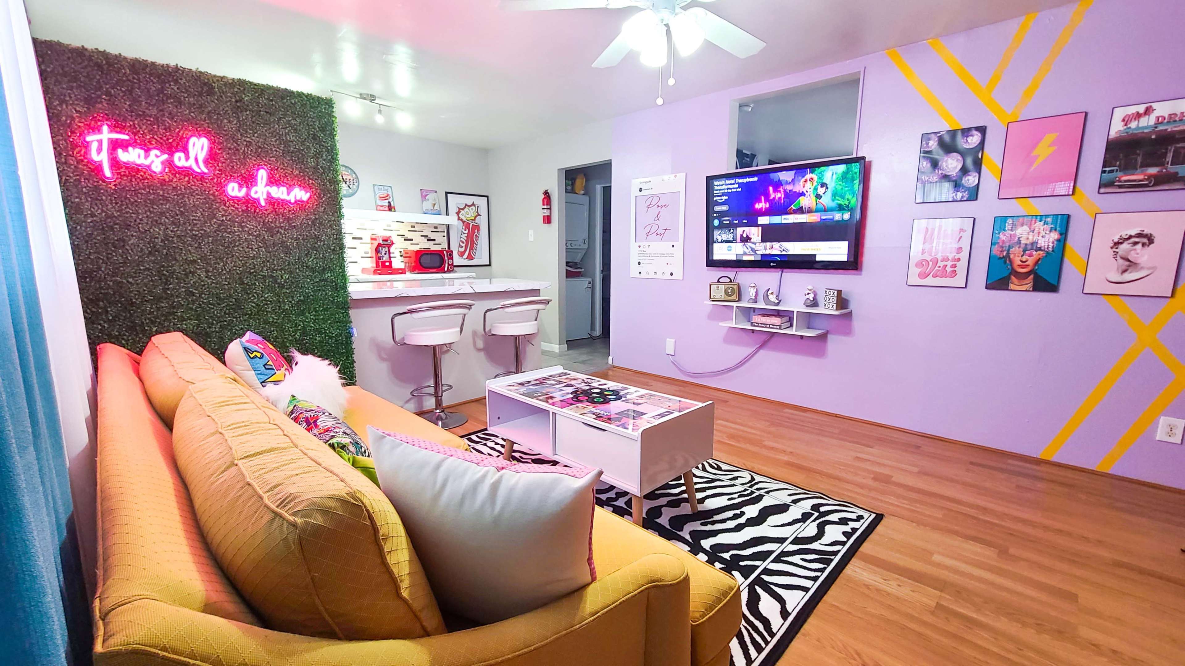 The image shows a brightly decorated living room with a yellow couch, a pink neon sign on a green wall, and a TV mounted on a purple wall adorned with framed art and a zebra-print rug.