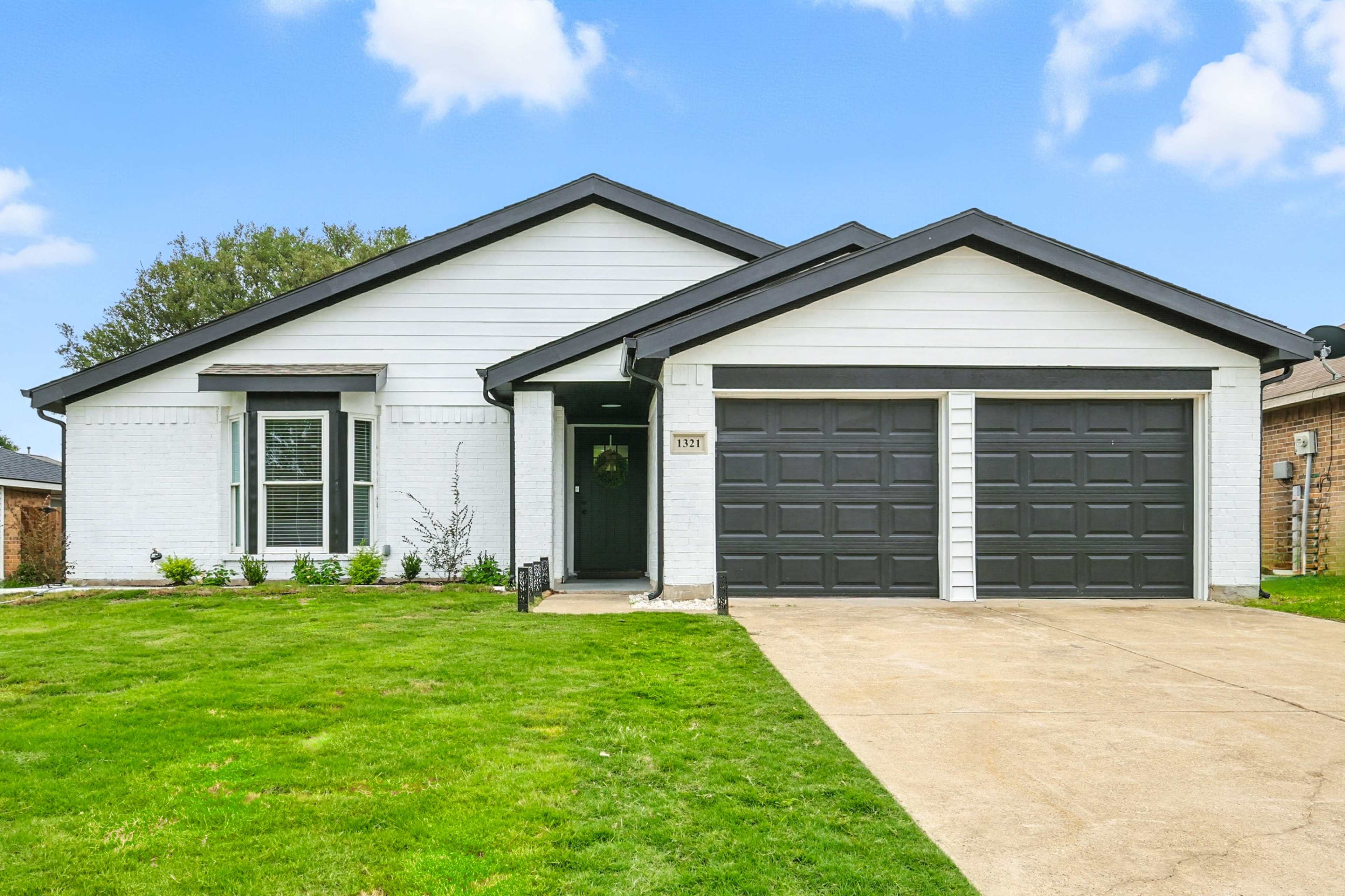 The image shows a single-story house with a white facade, black trim, and a two-car garage, situated on a well-maintained grassy lawn.