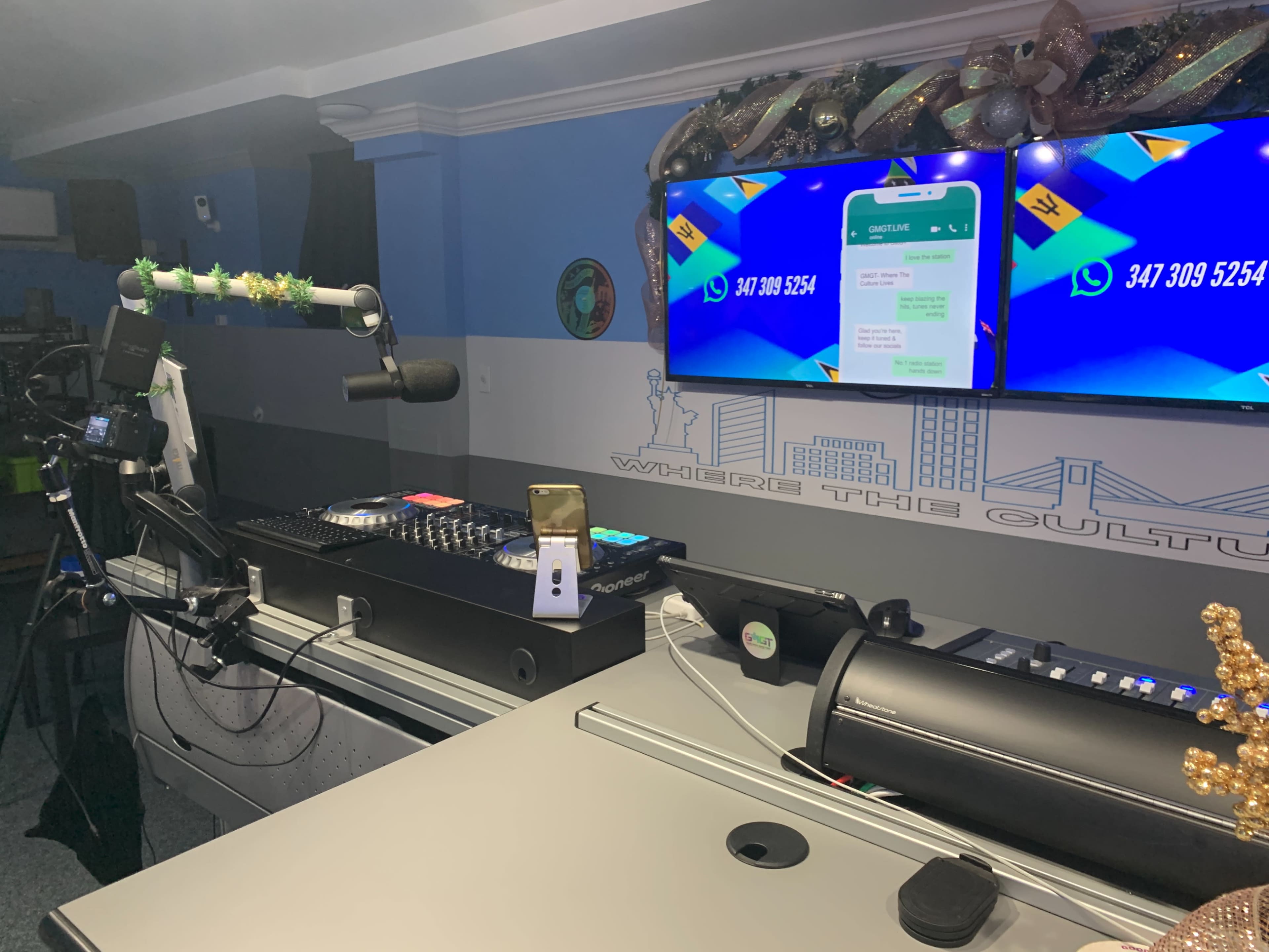 The image shows a broadcasting studio with a console, multiple screens displaying a messaging app, and a microphone setup.