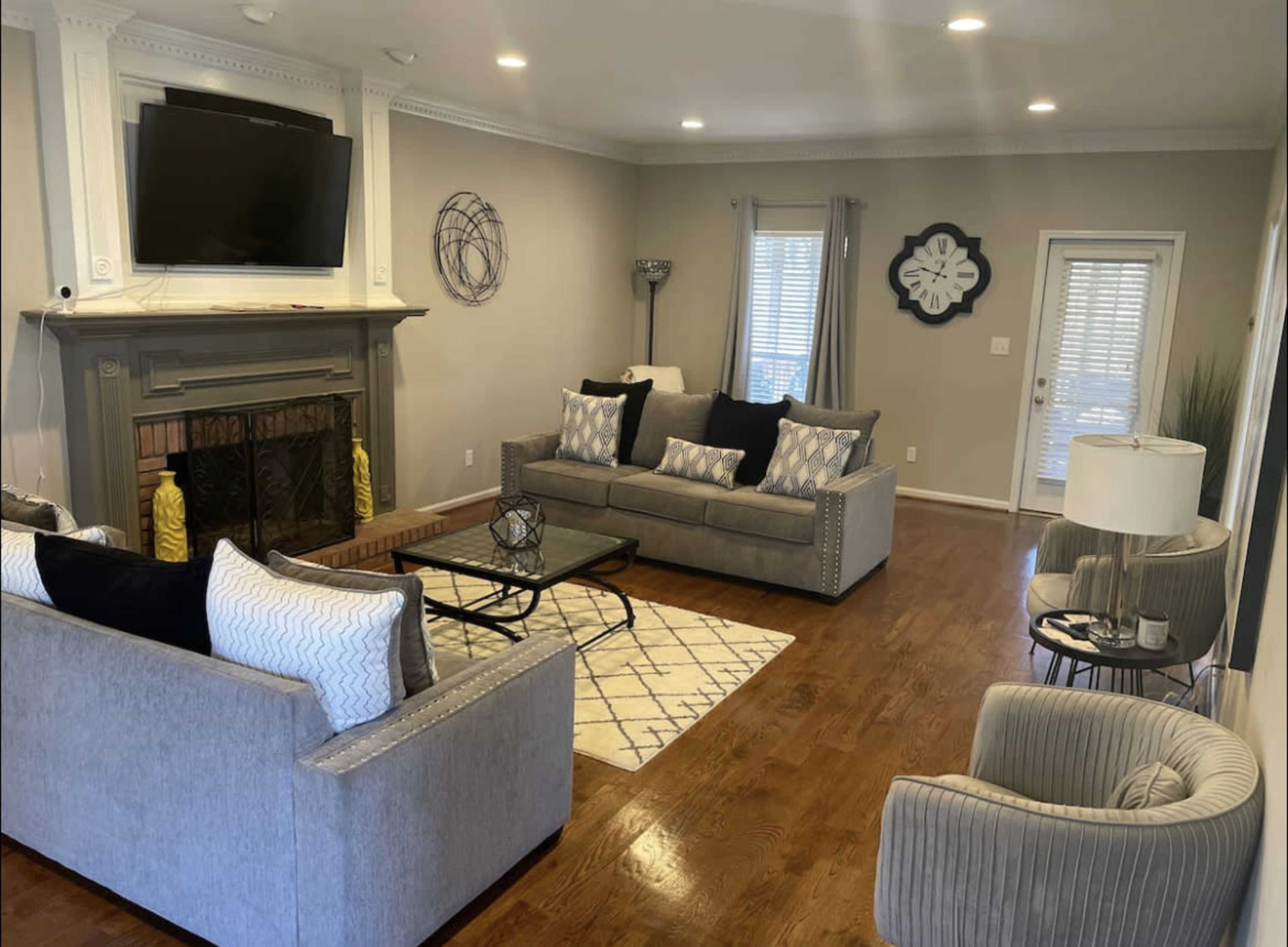 A living room features a gray sofa set, a coffee table on a patterned rug, a fireplace with a TV above it, and multiple windows providing natural light.