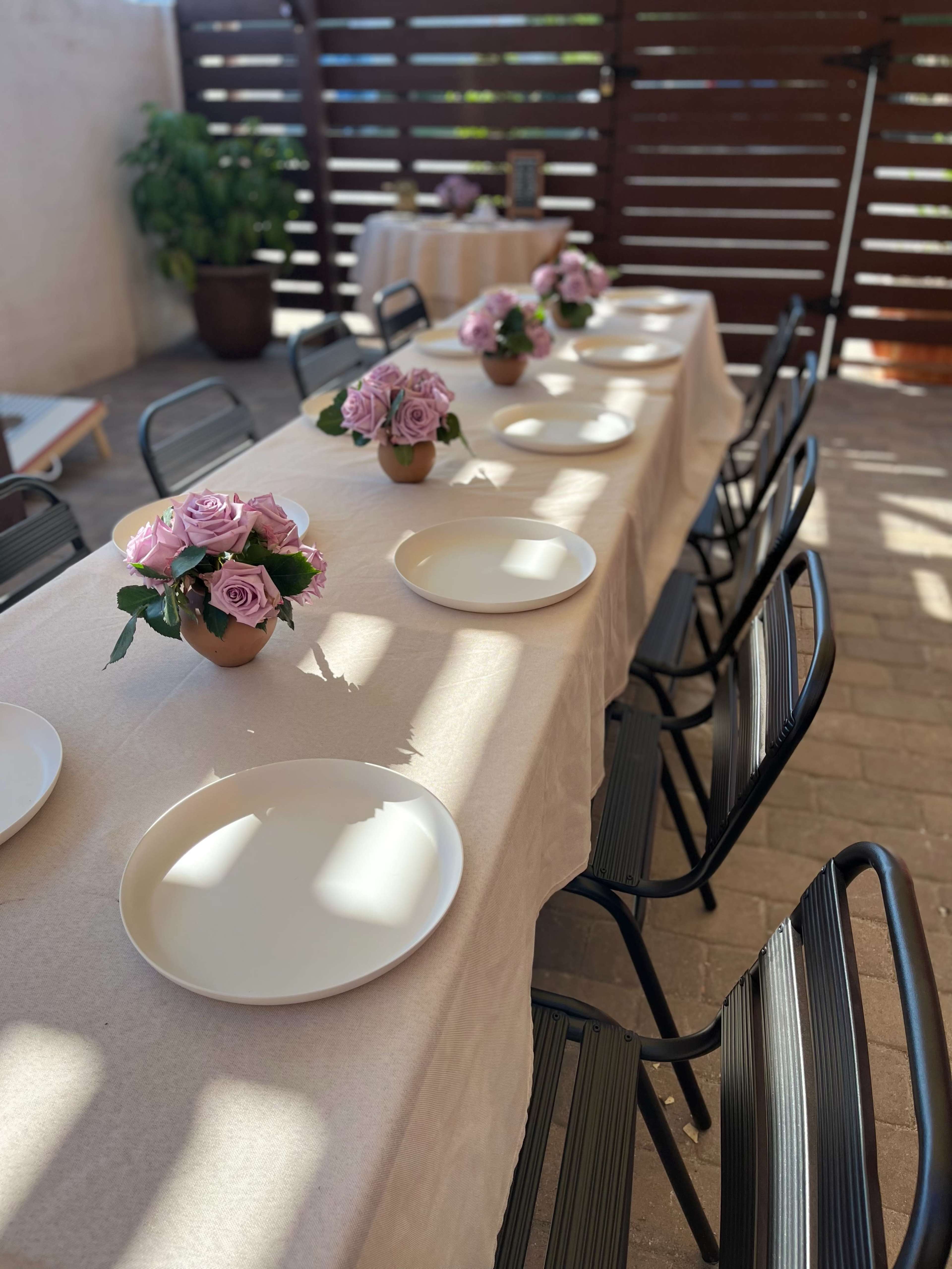 A long table is set outdoors with white plates and floral centerpieces, surrounded by black chairs under a wooden trellis.