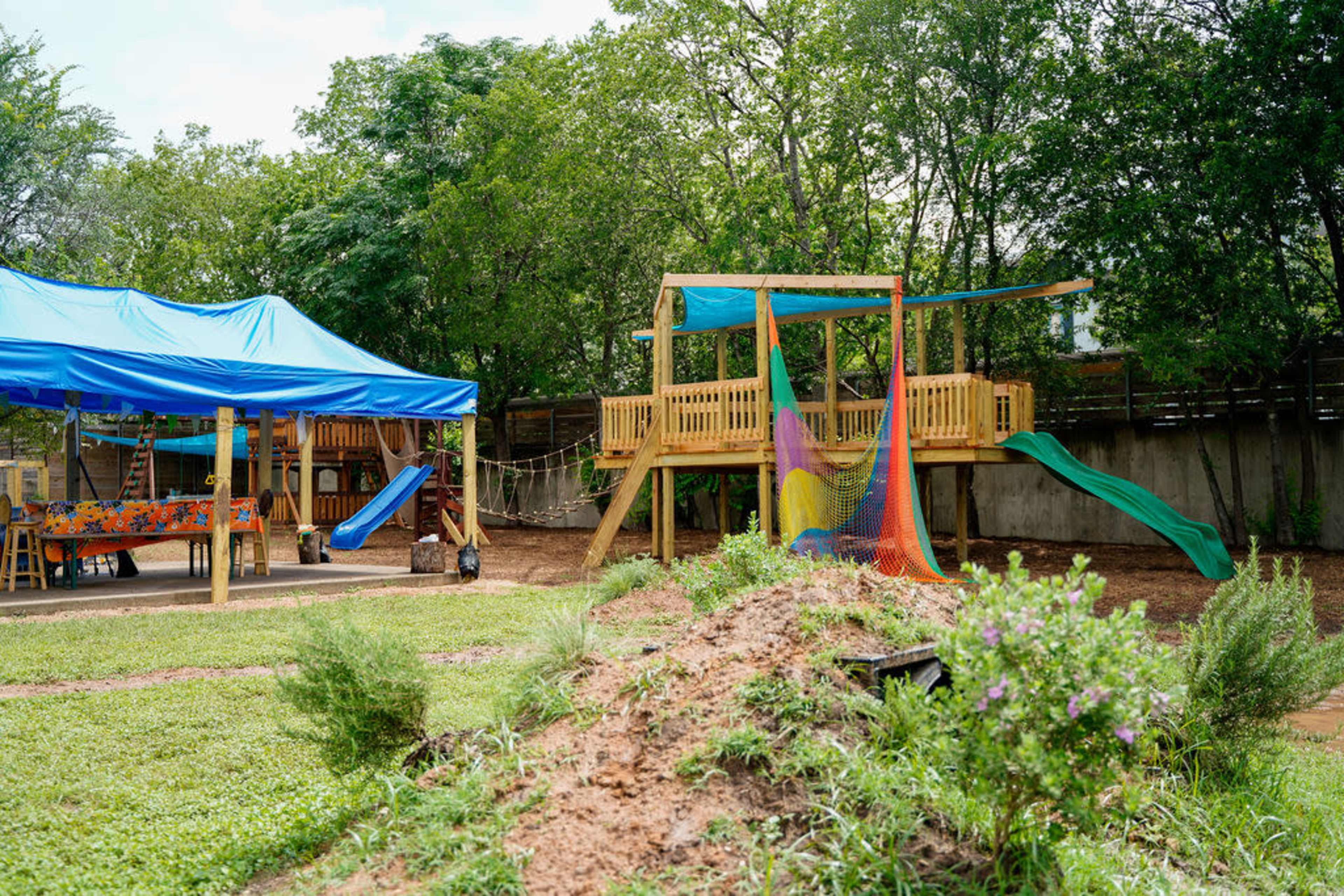 A wooden playground structure with slides and a climbing net is set up in a grassy area, adjacent to a shaded space featuring tables and chairs under a blue tent.