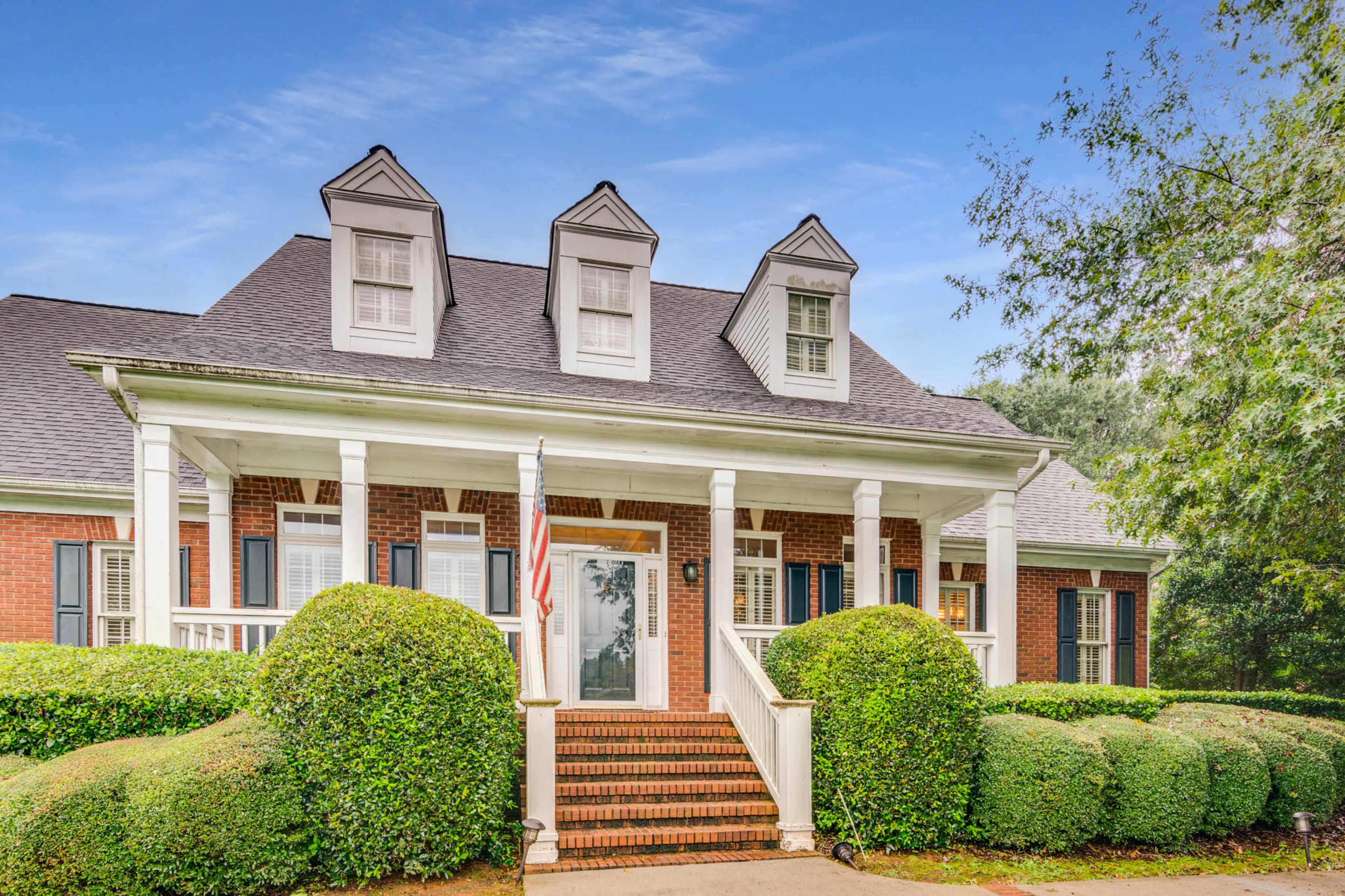 The image shows a two-story brick house with a front porch, surrounded by neatly trimmed bushes and a walkway leading to the entrance.