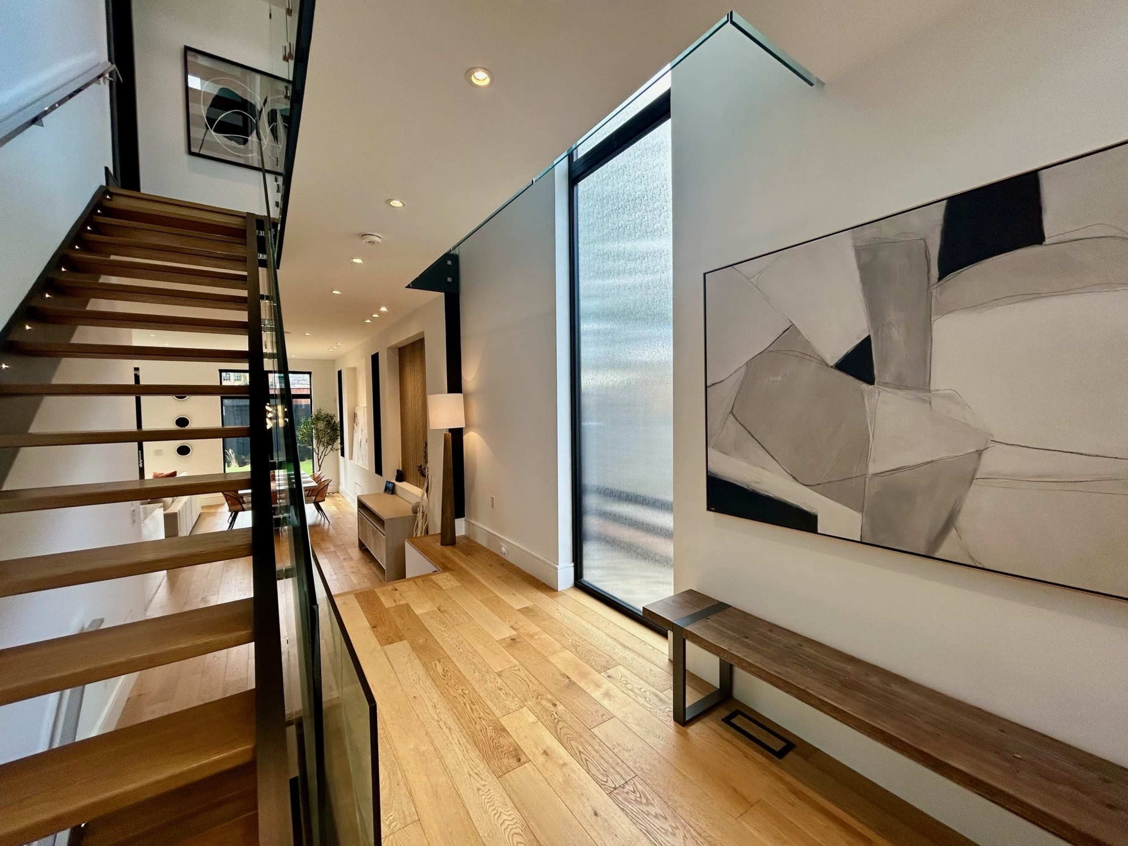 A modern interior space featuring a wooden staircase, a minimalist bench, and large windows that allow natural light to fill the area.