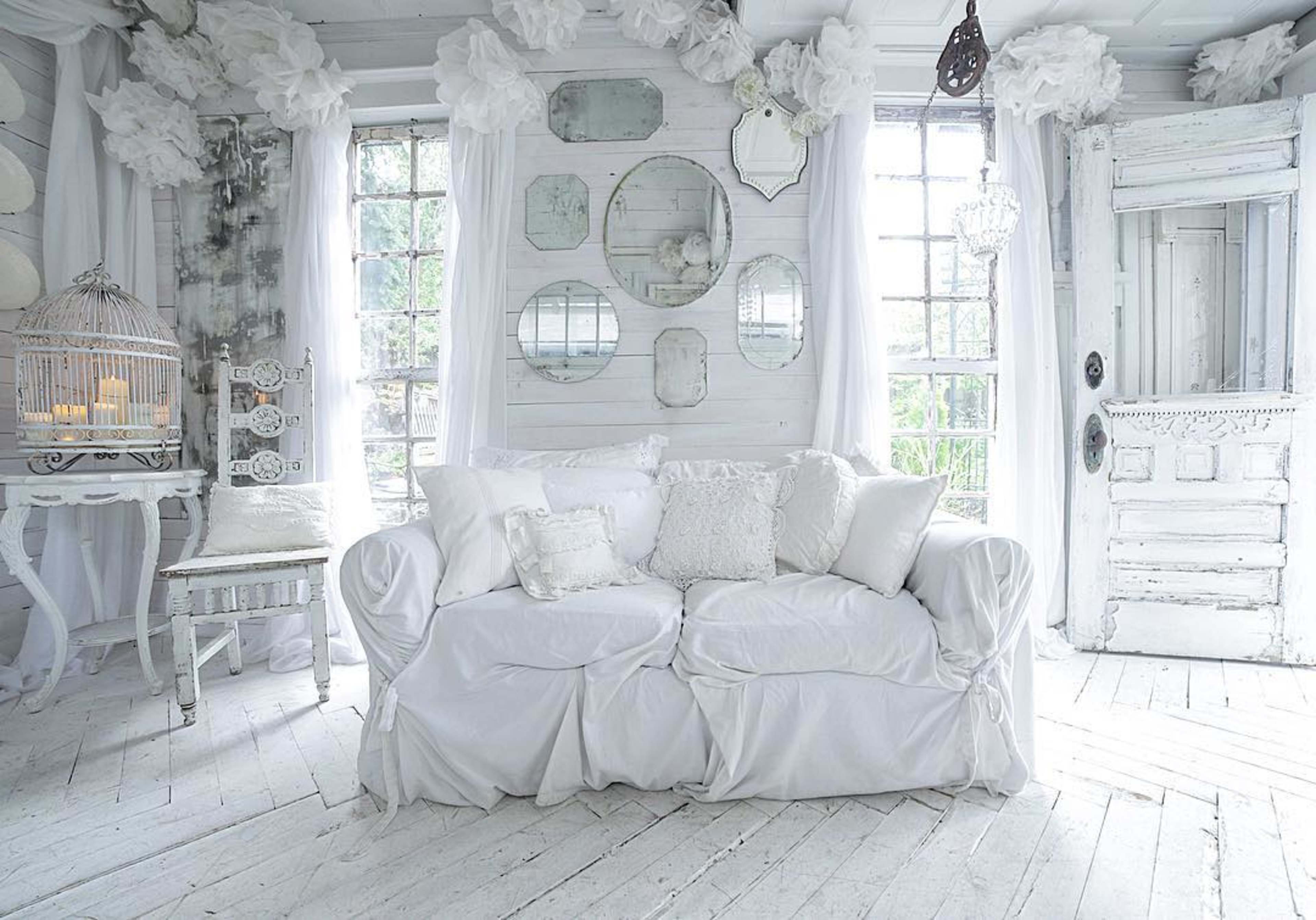 A bright, white living room features a large, draped couch, decorative mirrors on the wall, and vintage furnishings.