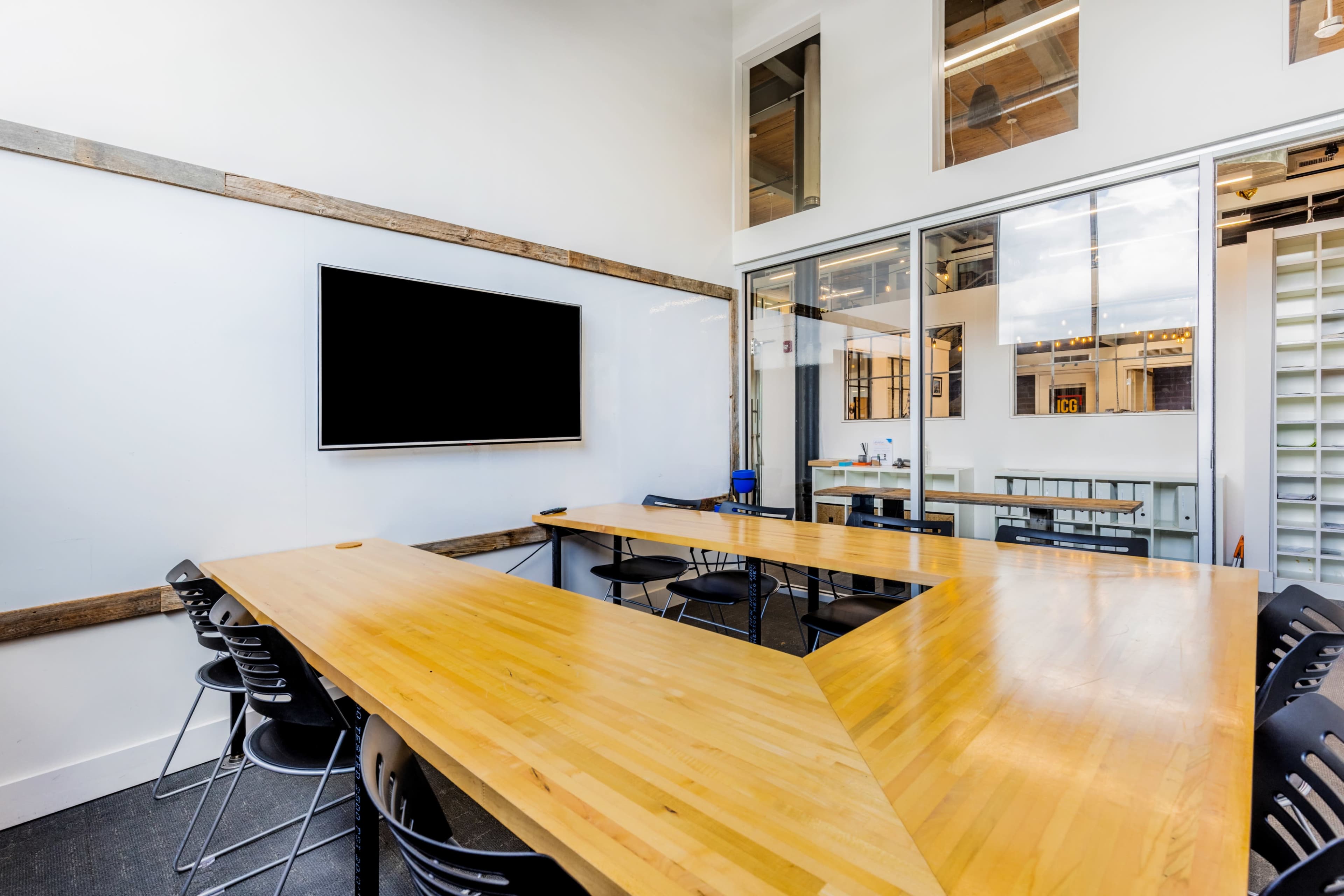 A modern conference room features a large wooden table arranged in a V-shape with black chairs and a wall-mounted screen.