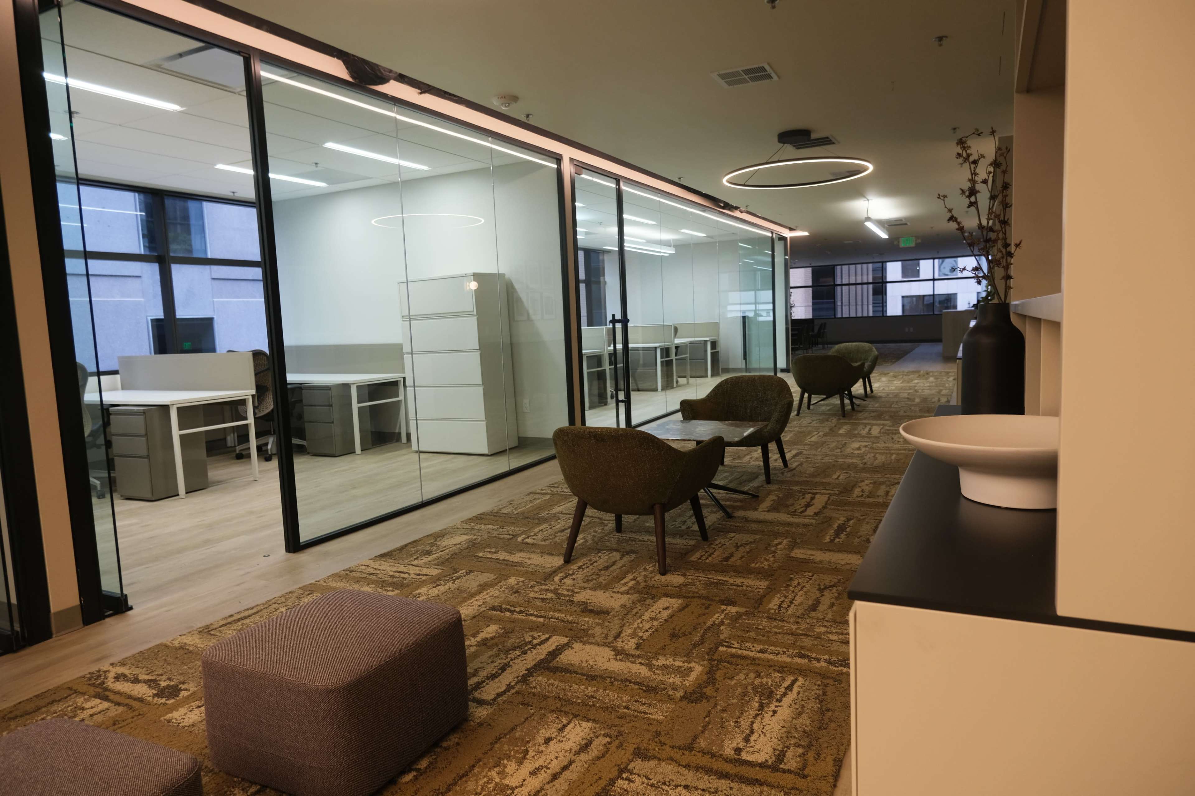A modern office interior with glass-walled meeting rooms, seating areas featuring chairs and ottomans, and a reception or common area.