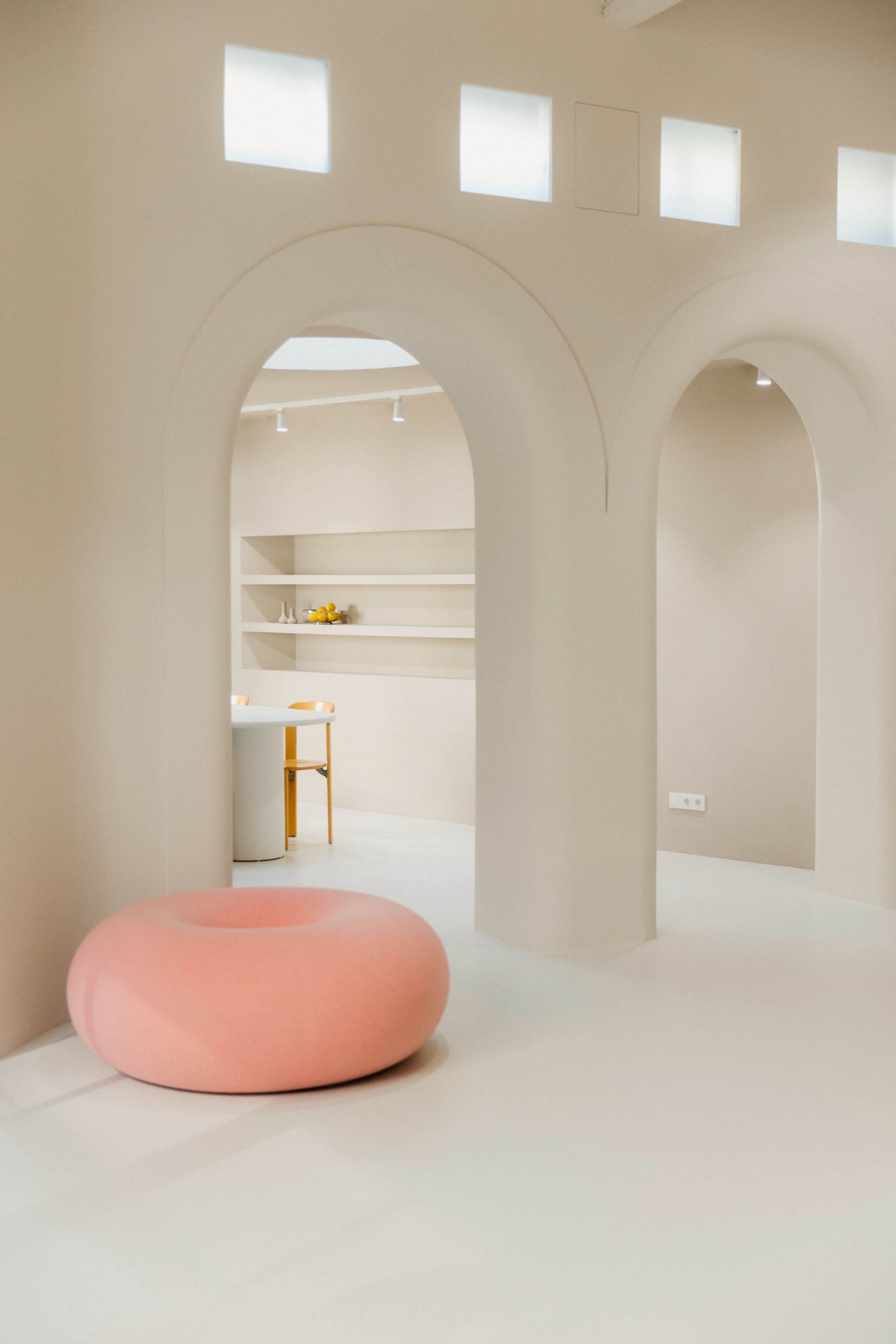 The image shows an interior space featuring arched doorways, a round pink seating element, and minimalist shelving in a softly lit environment.