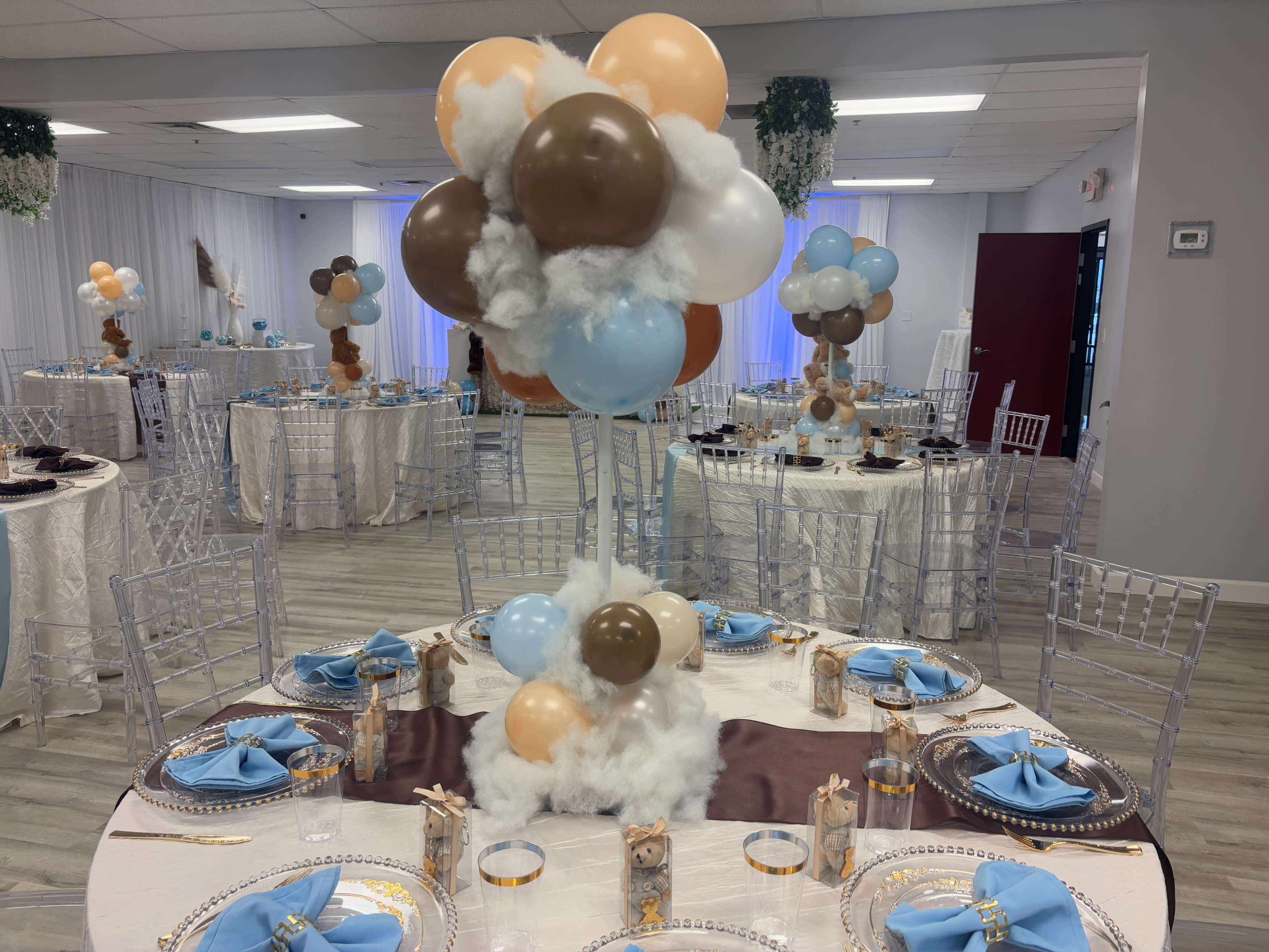 The image shows a decorated event space with several tables set for a celebration, featuring balloon centerpieces and neatly arranged tableware.