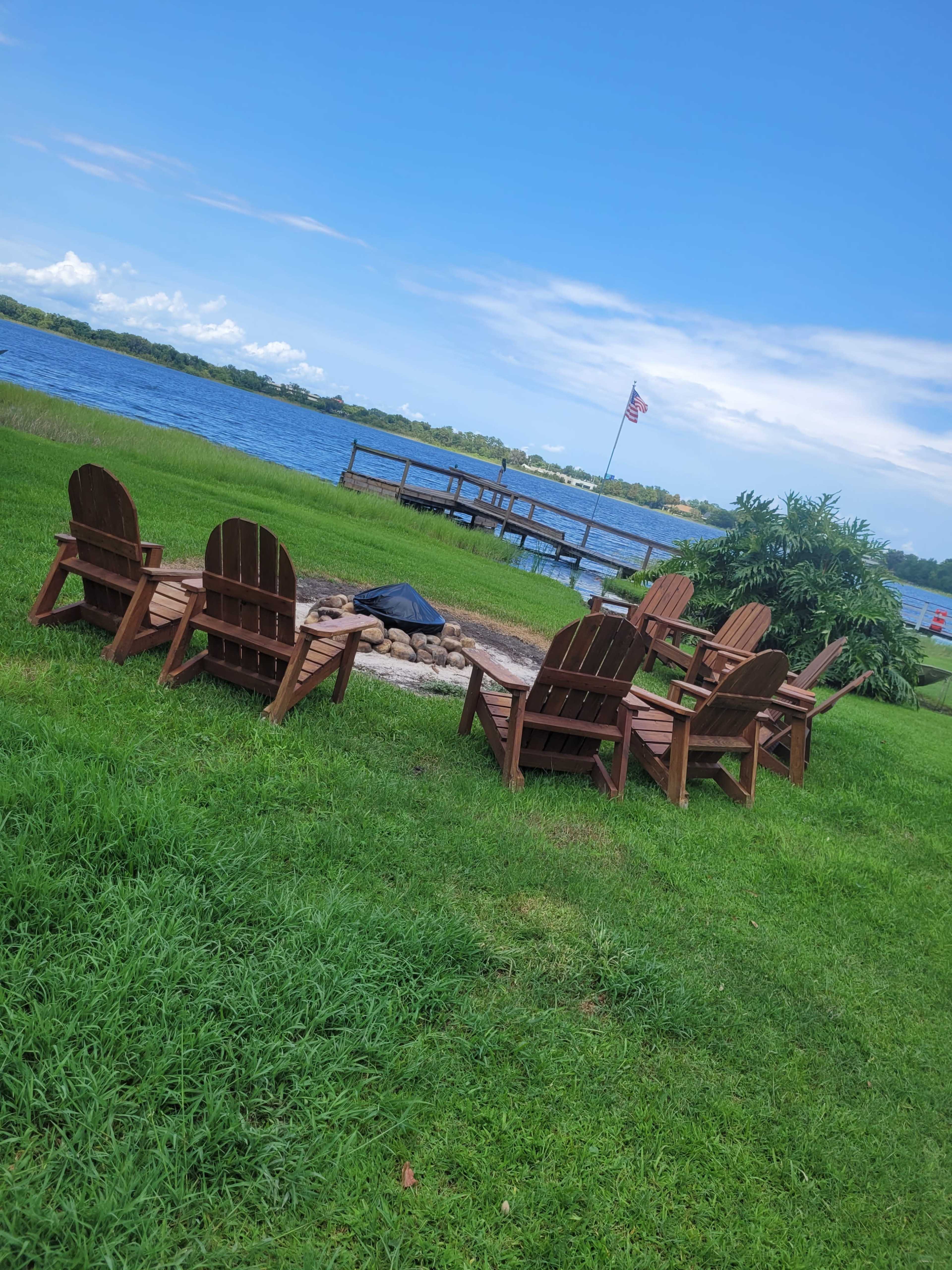 A circle of wooden chairs surrounds a fire pit near a lake, with a dock and an American flag in the background.