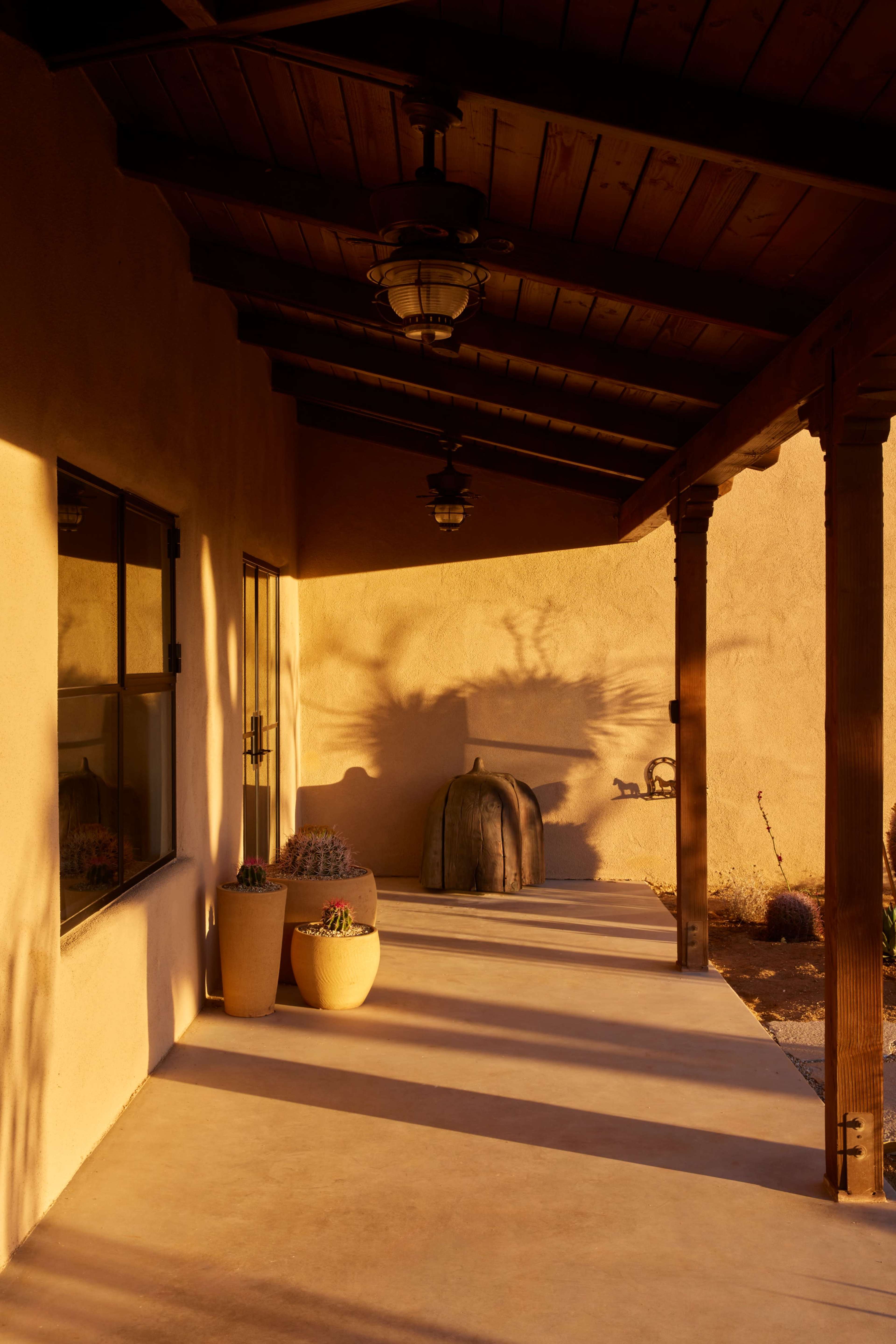 The image shows a beautifully lit outdoor porch area with potted plants and wooden beams casting shadows on the ground.