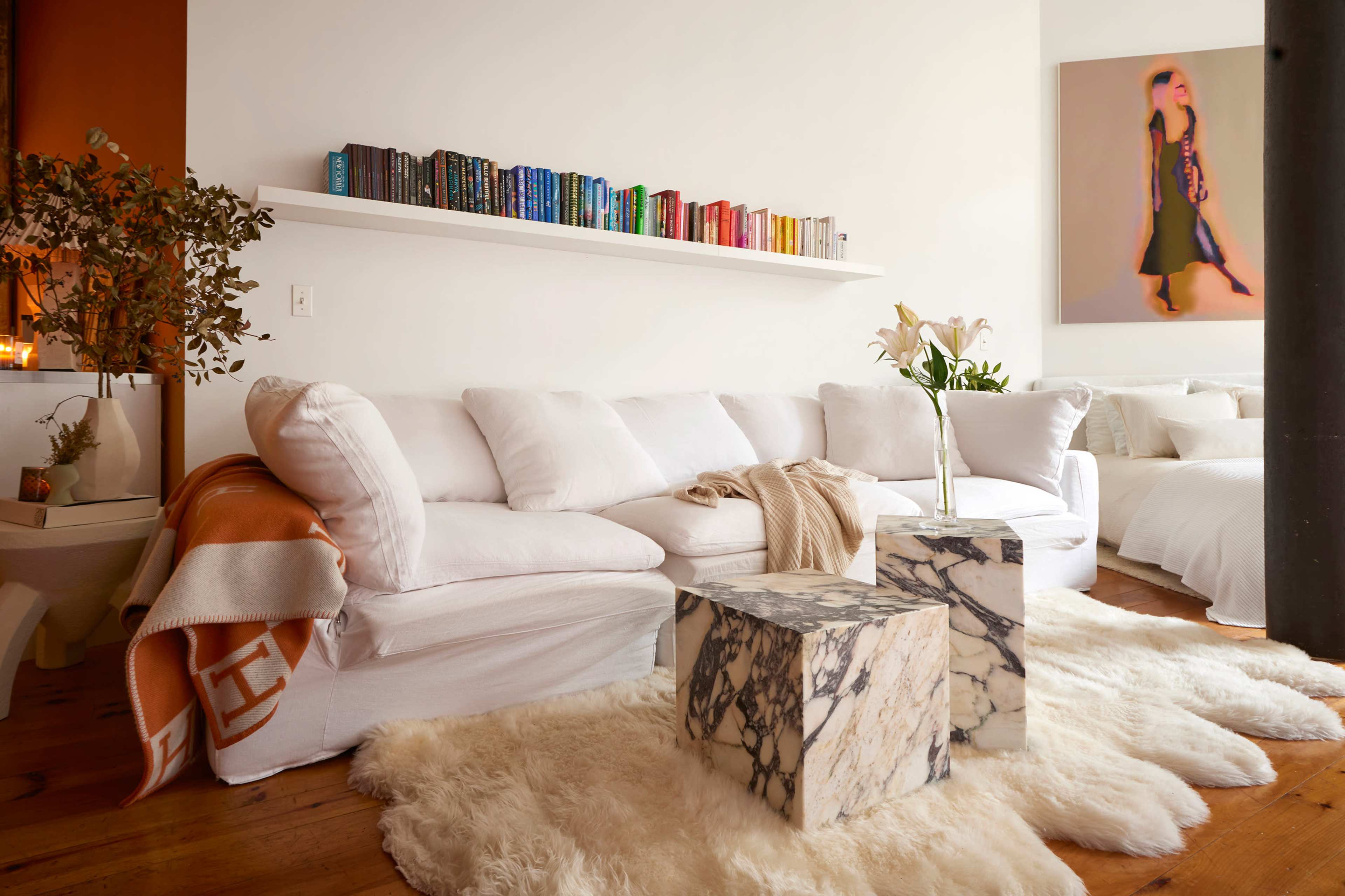 The image shows a cozy living room featuring a white sectional sofa, a textured area rug, and two marble side tables, complemented by a bookshelf filled with colorful books above.