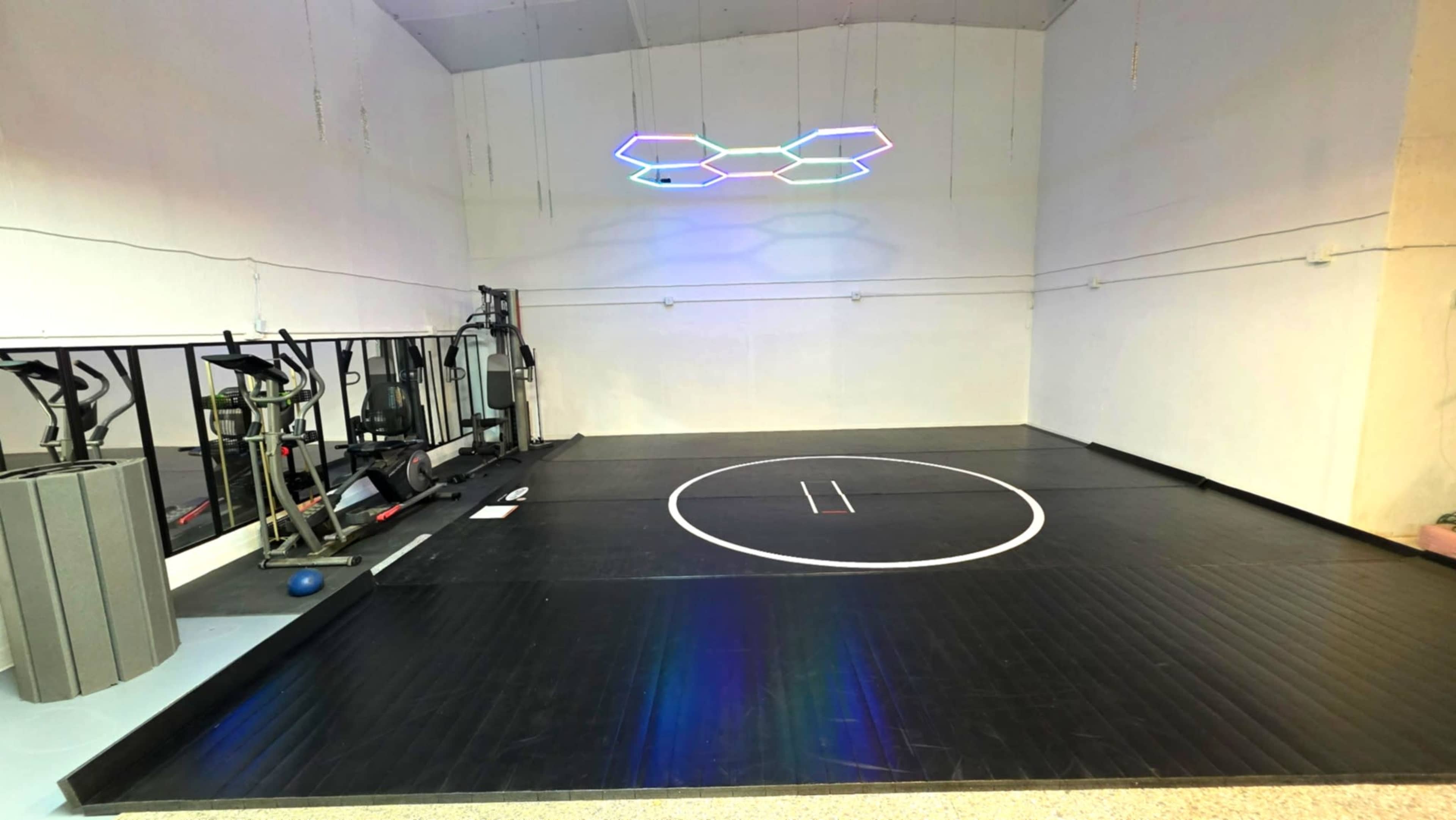 The image shows a spacious indoor gym with a wrestling mat in the center, exercise equipment along one wall, and colorful LED lighting on the ceiling.