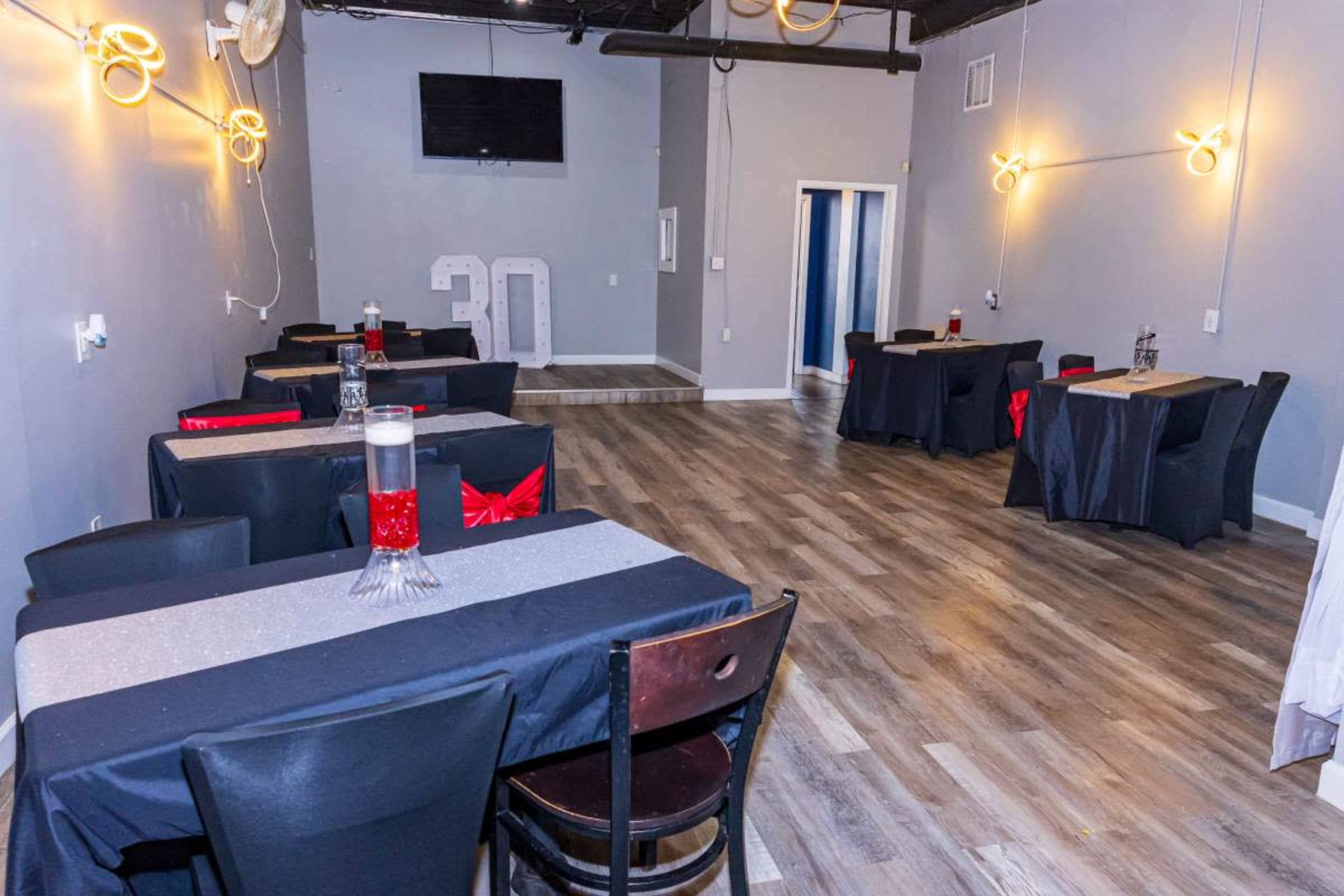 The image shows a spacious event room with black tables covered in dark tablecloths and decorated with red accents, featuring a television on the wall and large numerals "30" displayed.