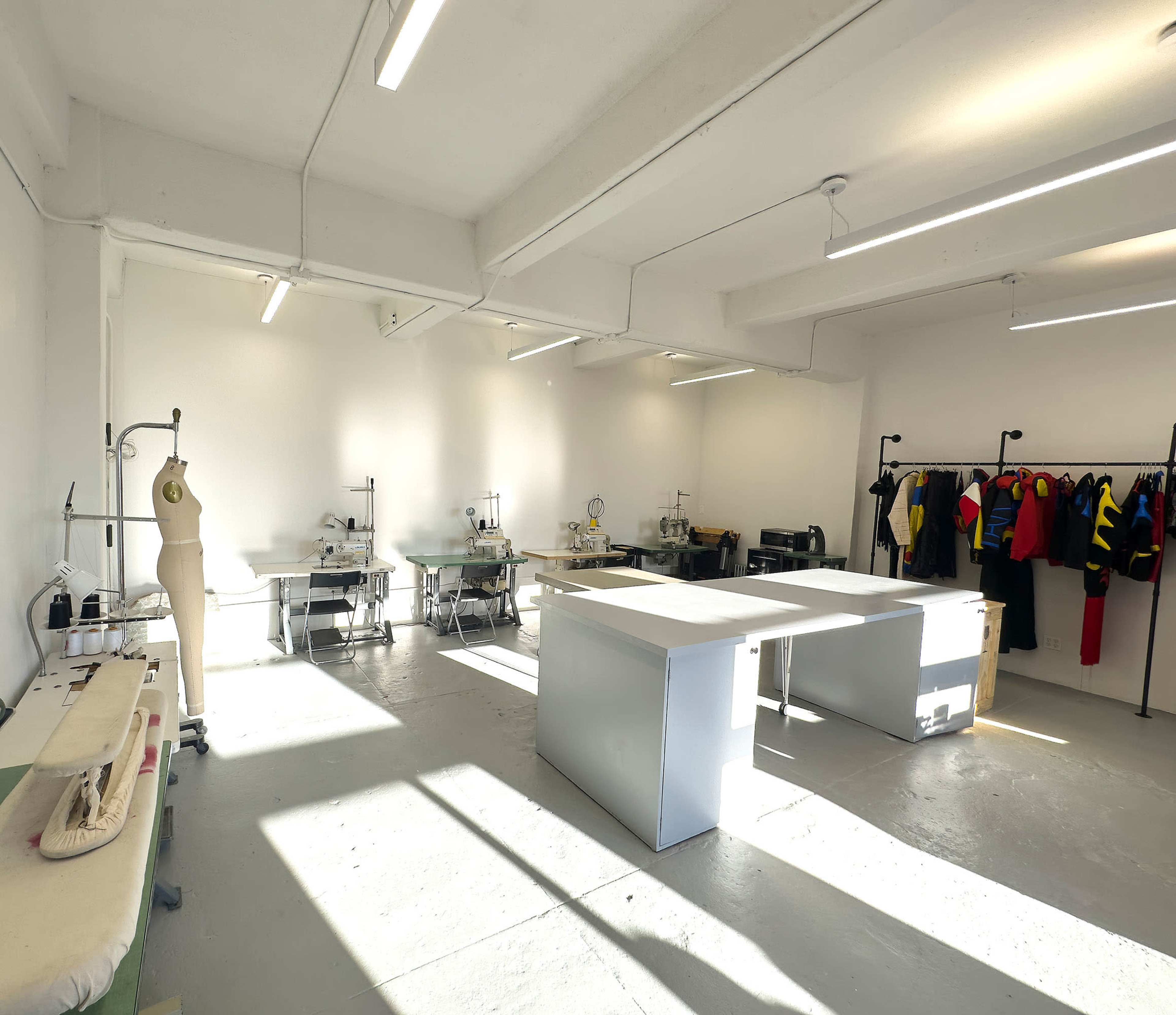 A spacious, well-lit textile studio features work tables, sewing machines, and a clothing rack with various garments.