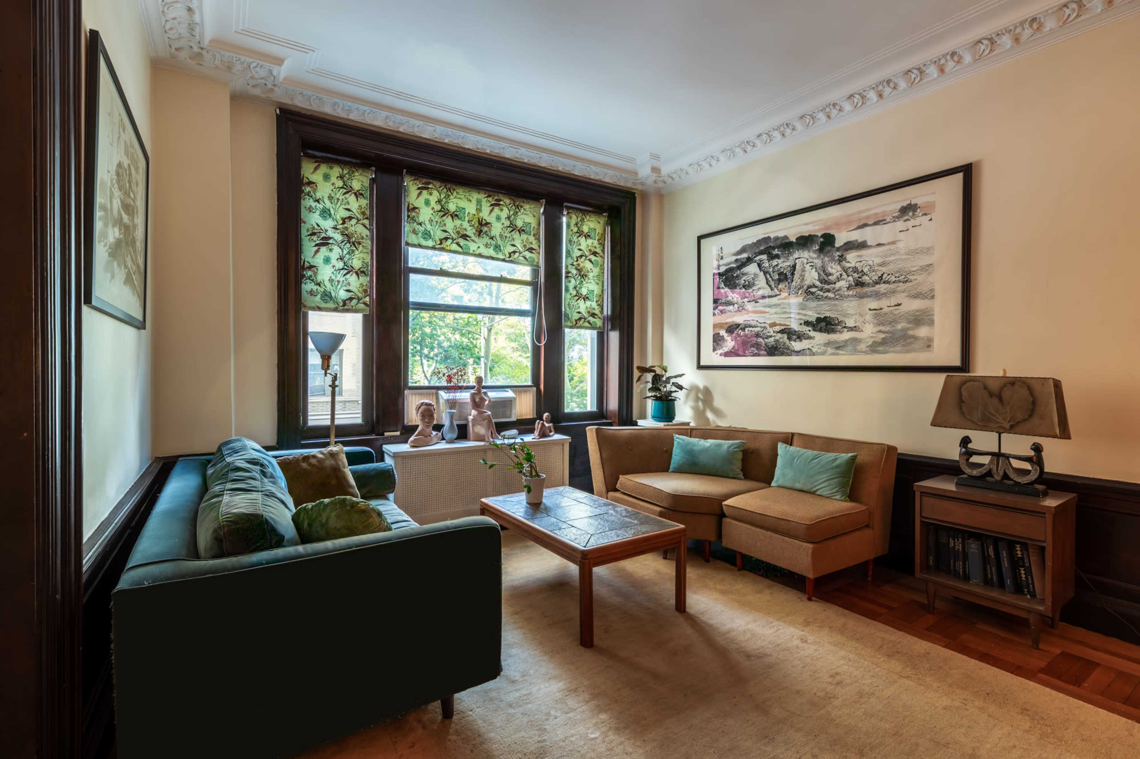 The image shows a well-furnished living room with large windows, green and beige furniture, and a decorative painting on the wall.