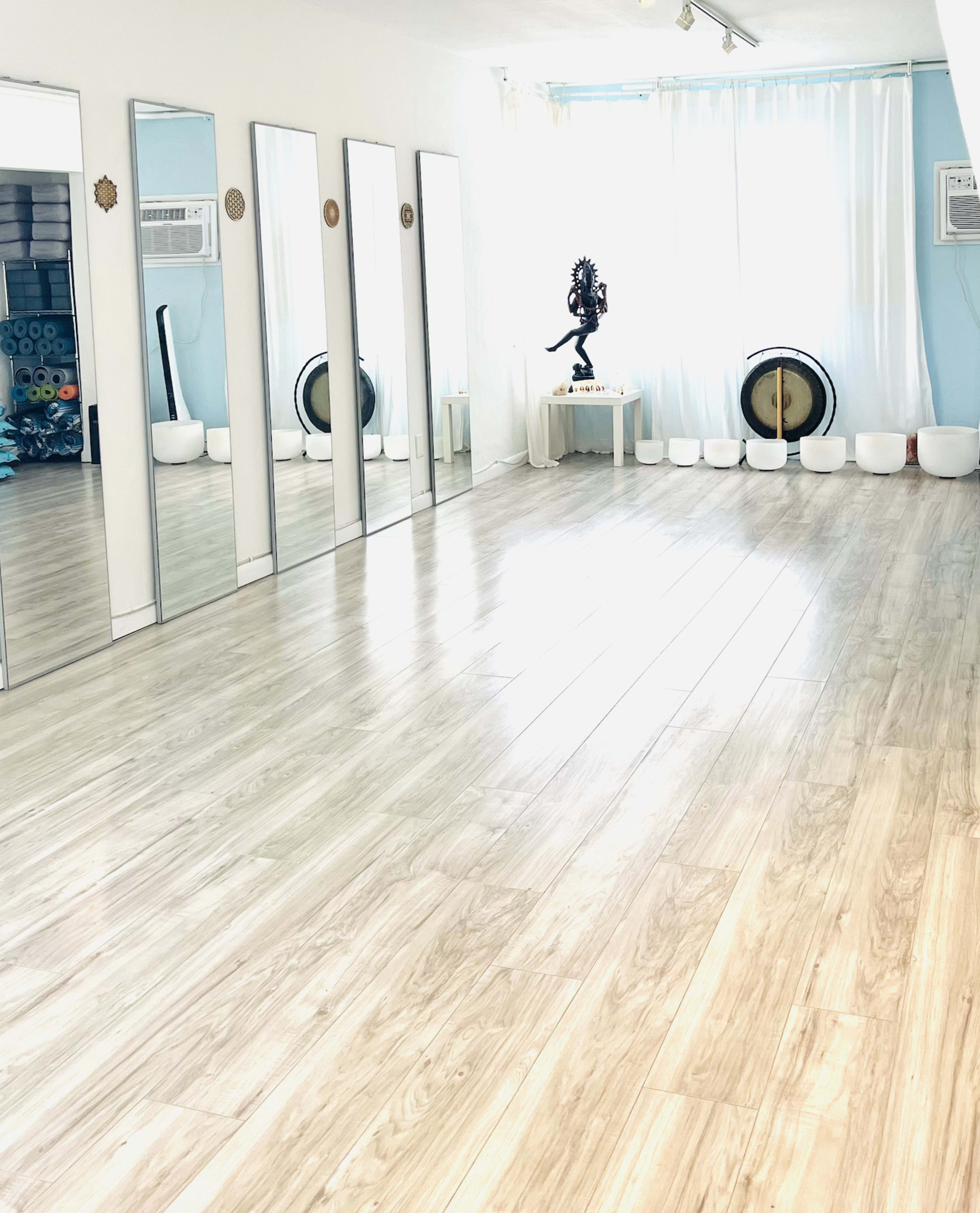 The image shows a spacious, well-lit room with mirrors along one wall, wooden flooring, and a few decorative items, including a statue and musical instruments.