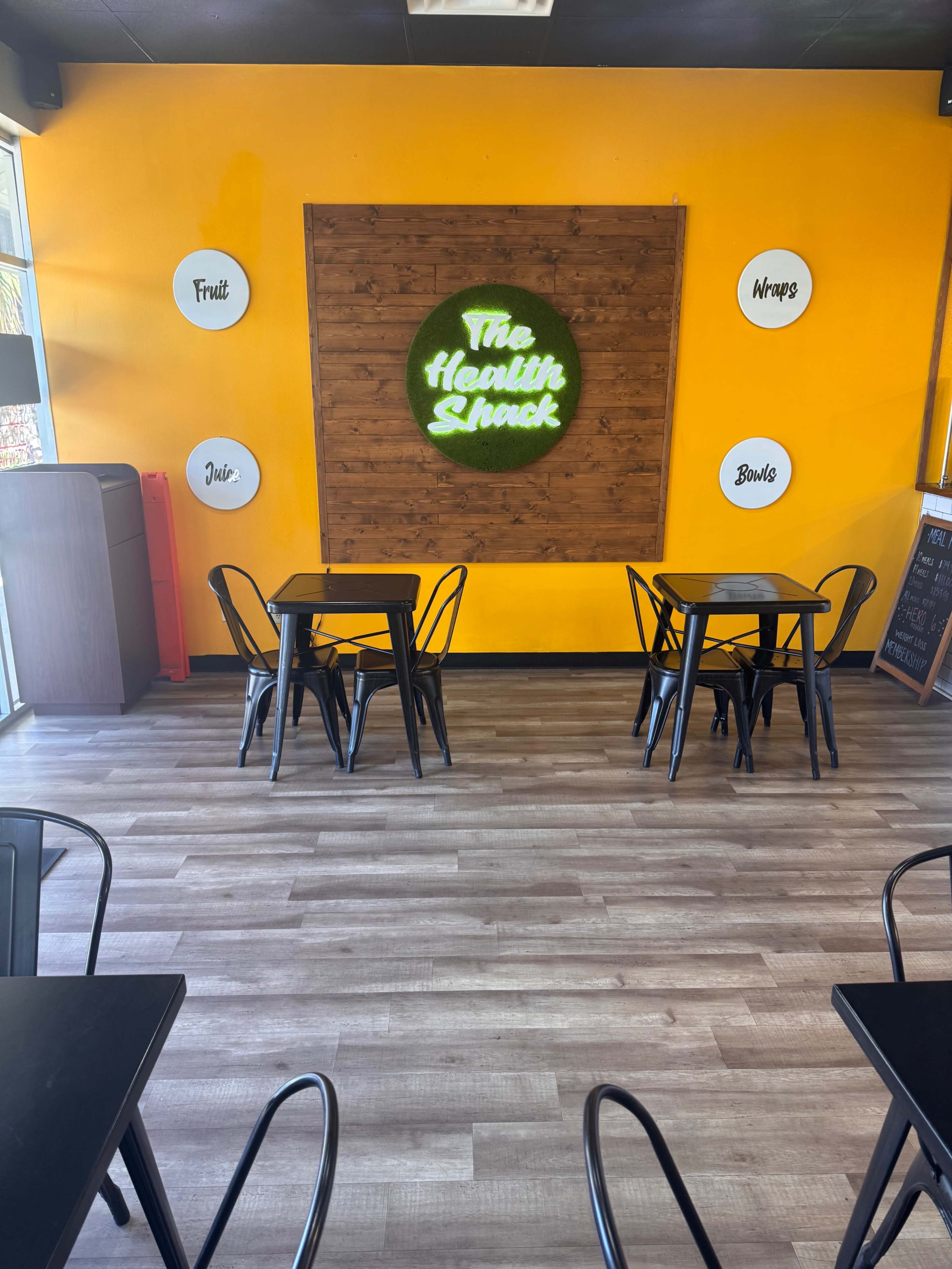 The interior of a small cafe features black tables and metal chairs against a bright yellow wall with a wooden accent displaying a circular green sign that reads "The Health Shack."