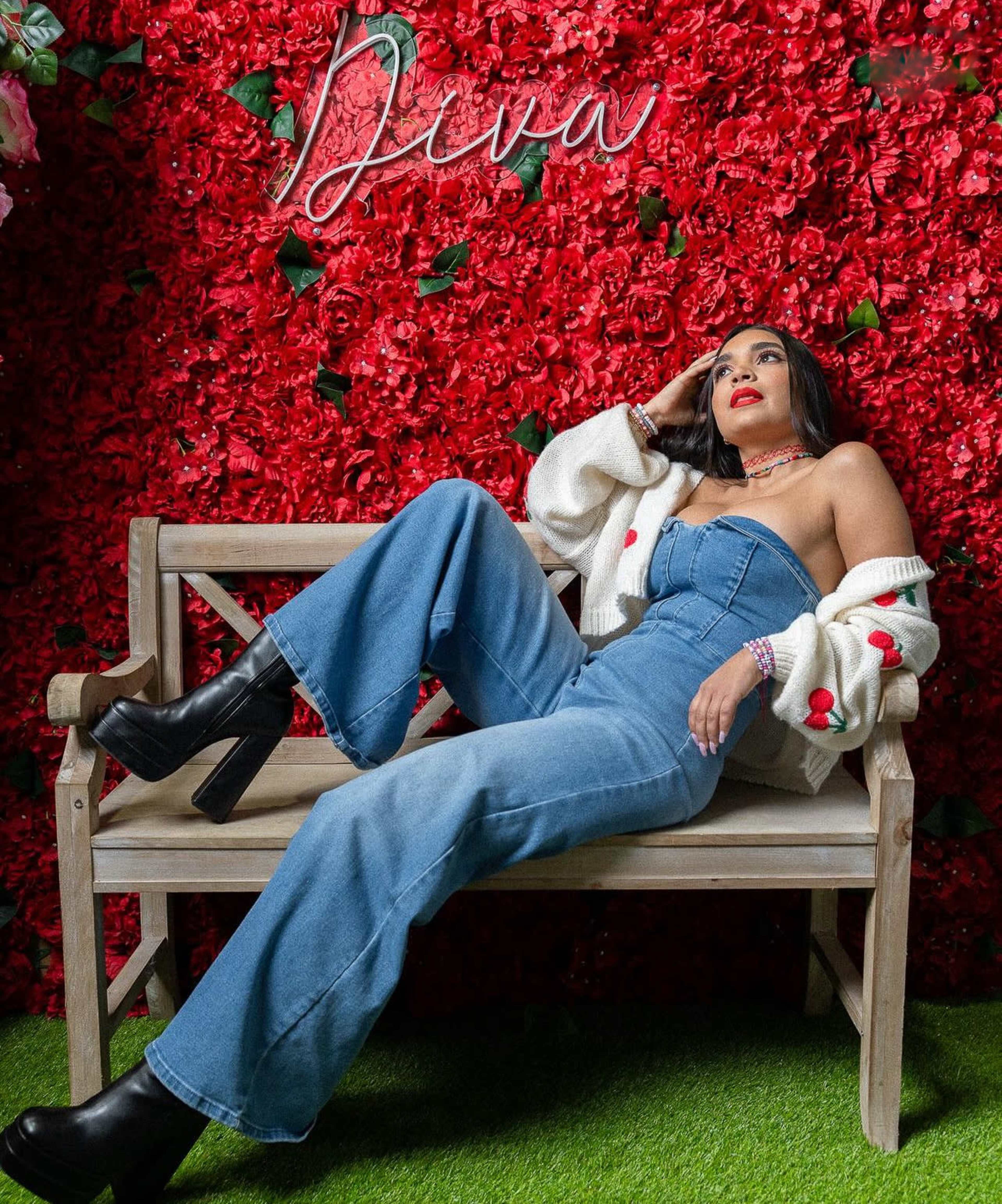 A person poses on a wooden bench in front of a wall covered in red flowers, wearing a strapless denim top and flared jeans.