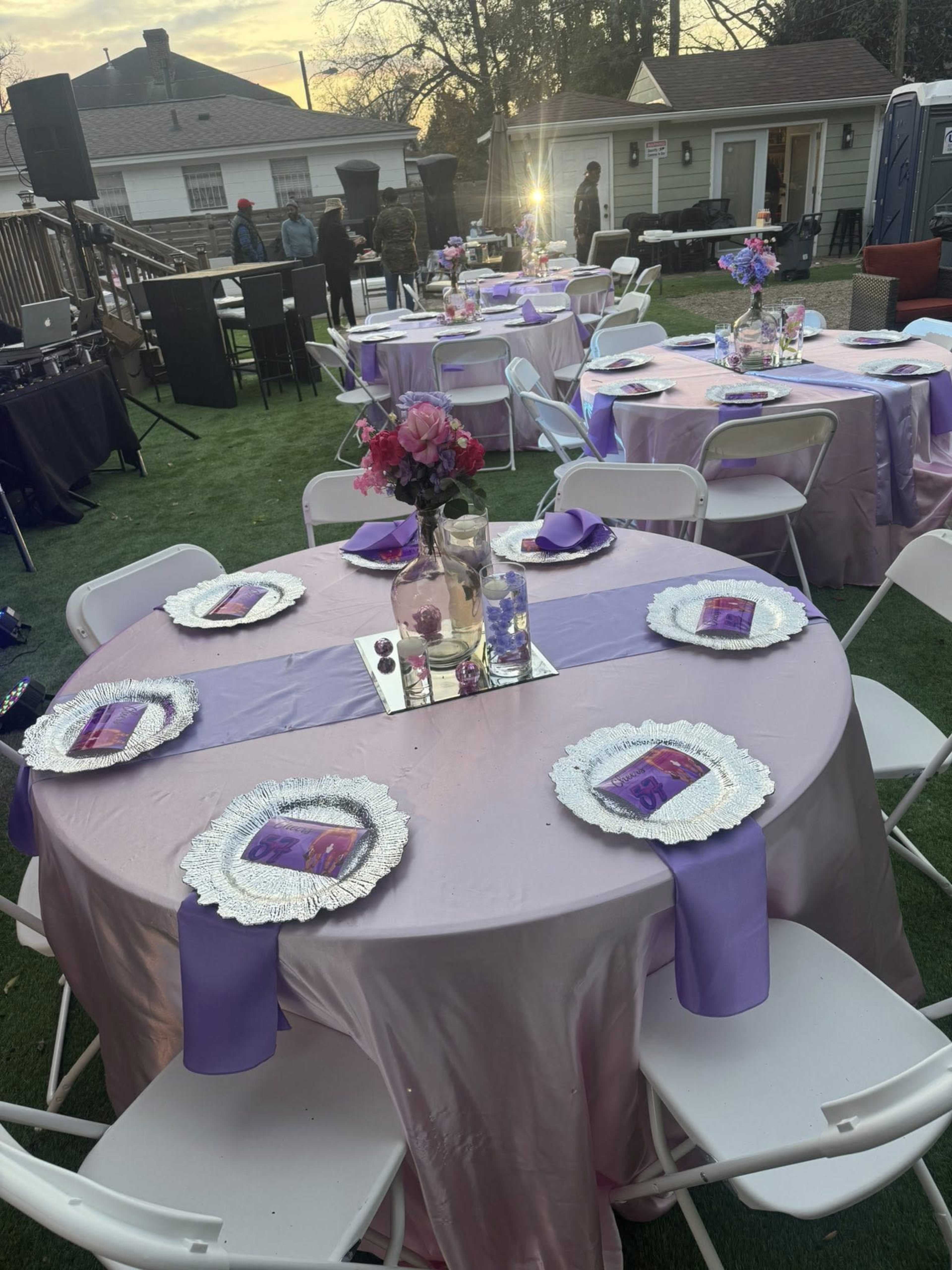 The image shows a set of elegantly arranged tables with pink tablecloths, each featuring decorative centerpieces and place settings, in a yard during an outdoor event.