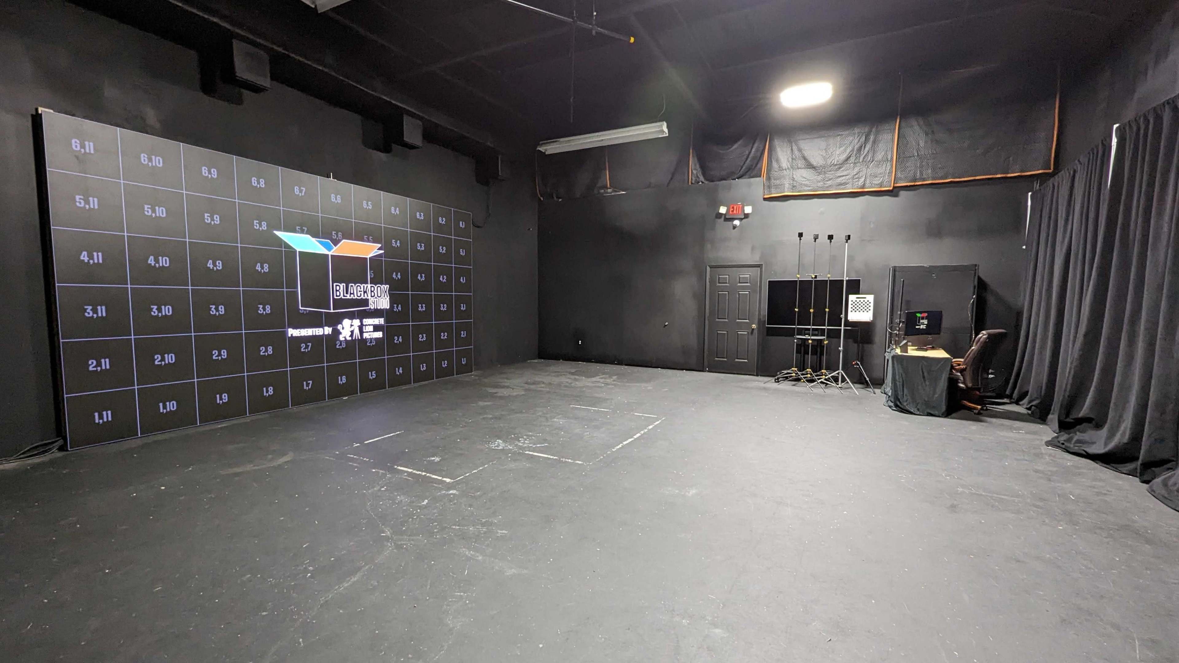 A large, empty black-walled studio equipped with a grid display panel on one side and a control area with electronic equipment in the corner.
