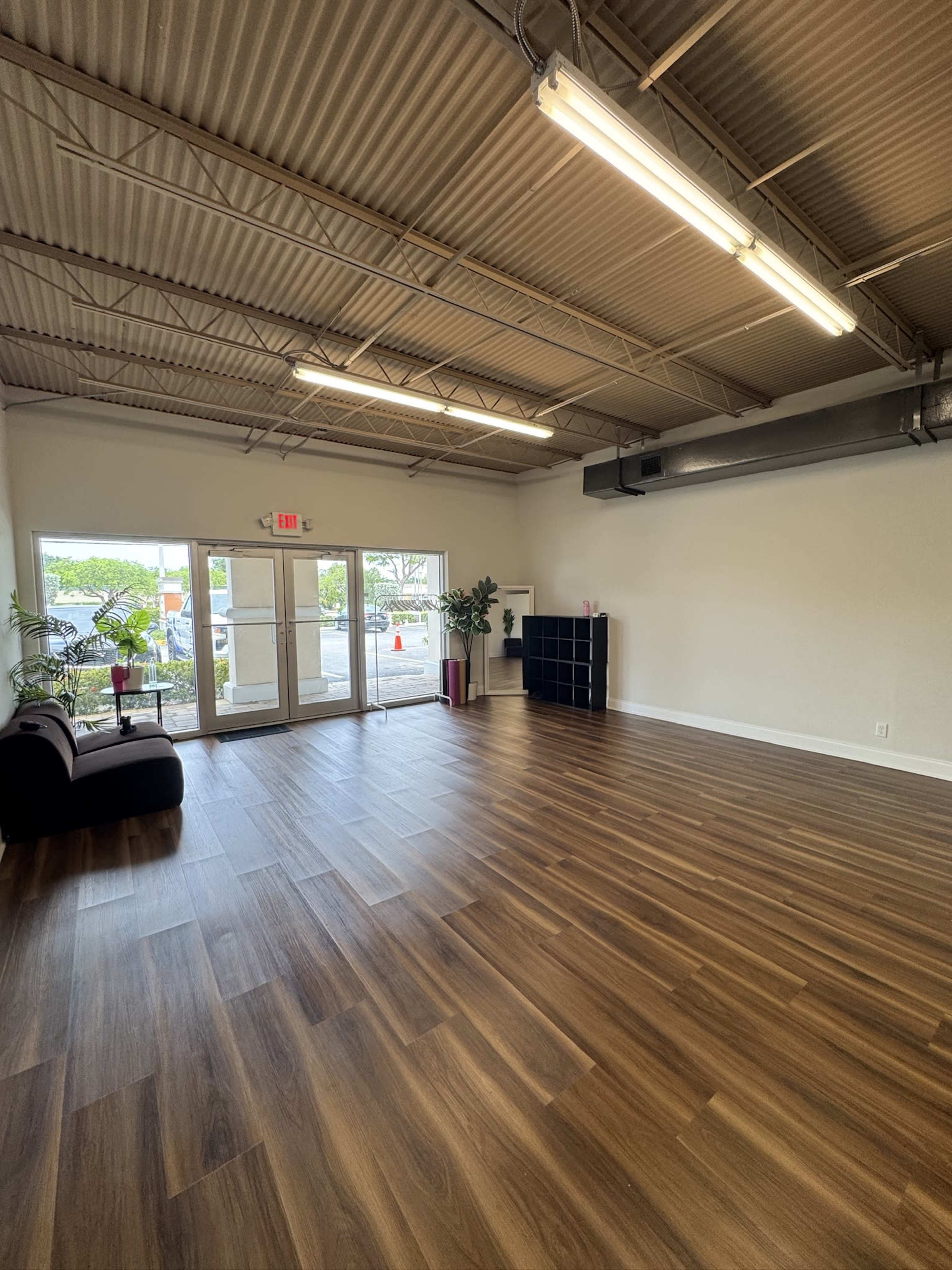 The image shows a spacious, empty room with wooden flooring, large front windows, and minimal furnishings, including plants and a bookshelf.