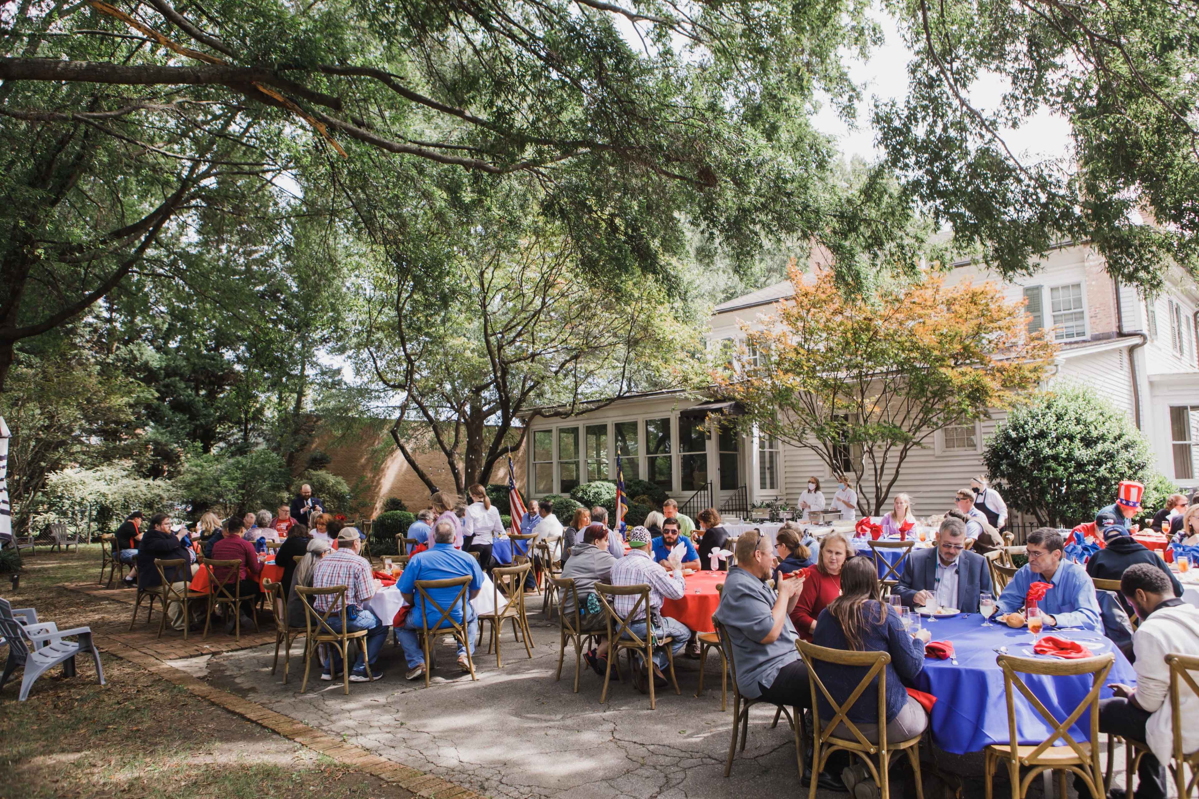 A gathering of people sitting at tables covered with red and blue tablecloths in a shaded outdoor setting near a house.
