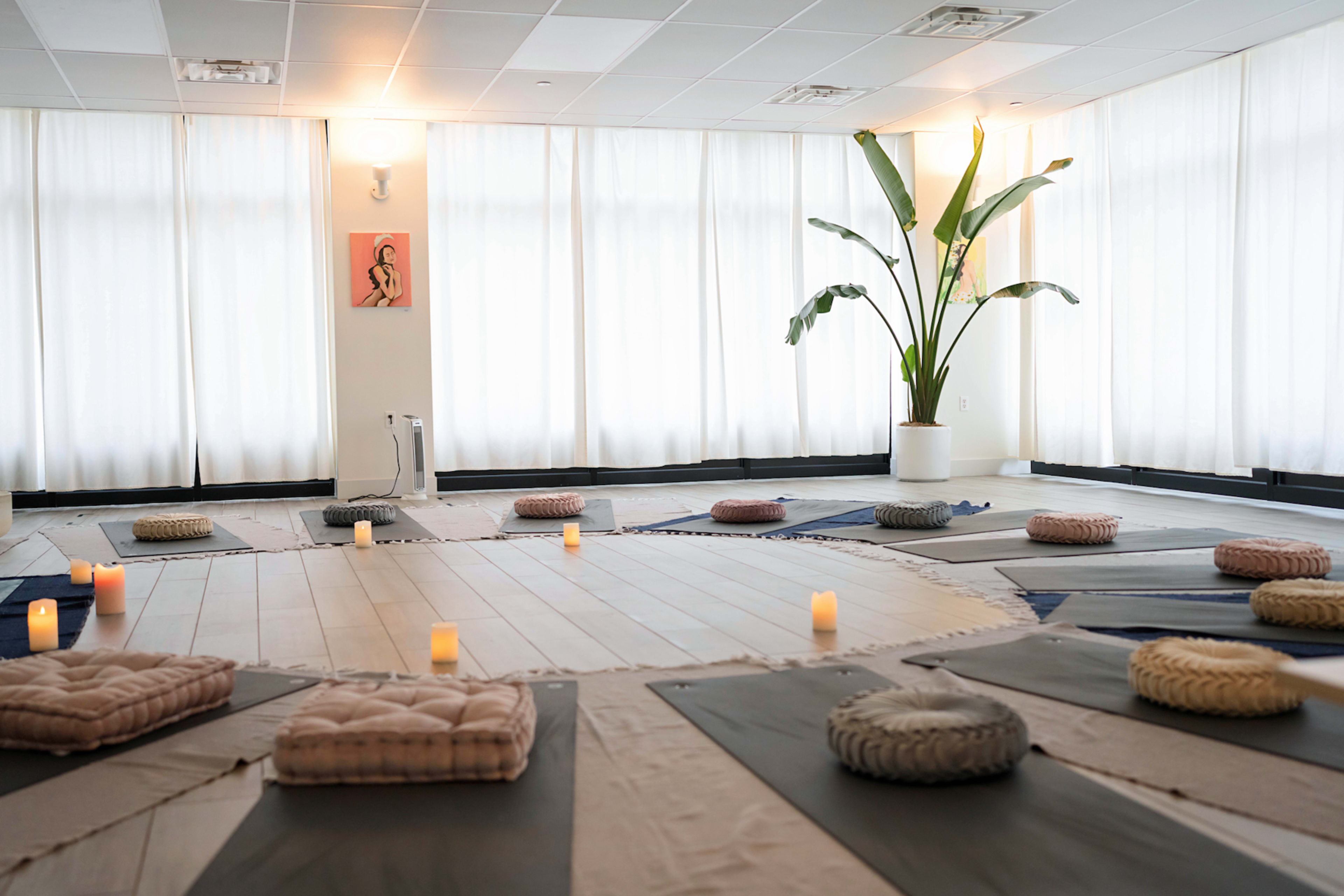 A spacious meditation room arranged with floor cushions and candles, surrounded by large windows draped with white curtains and a potted plant in the corner.