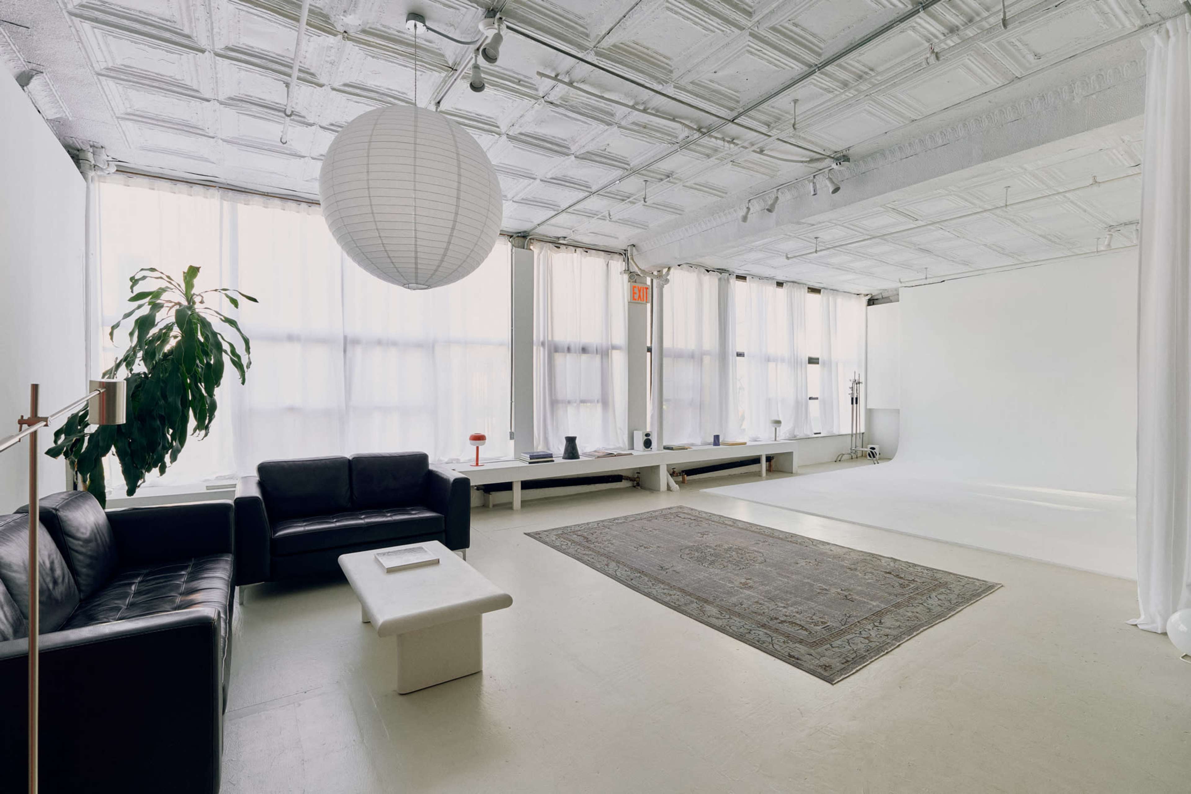 A minimalist studio space features a black leather sofa, a light-colored coffee table, and a large area rug on a polished floor, with ample natural light coming through sheer curtains.