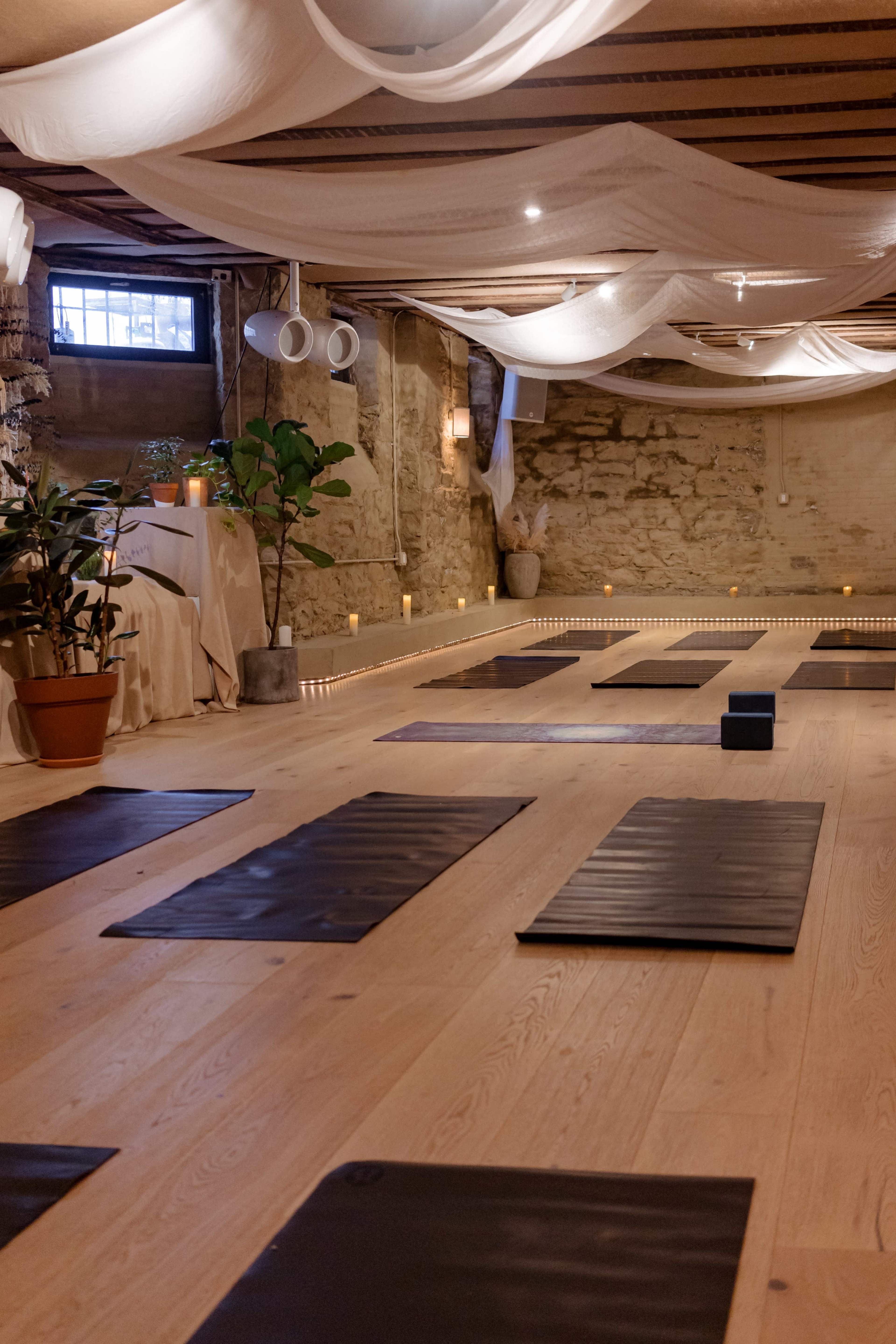 The image shows a serene yoga studio with several yoga mats arranged on a wooden floor, surrounded by candles and soft drapery.