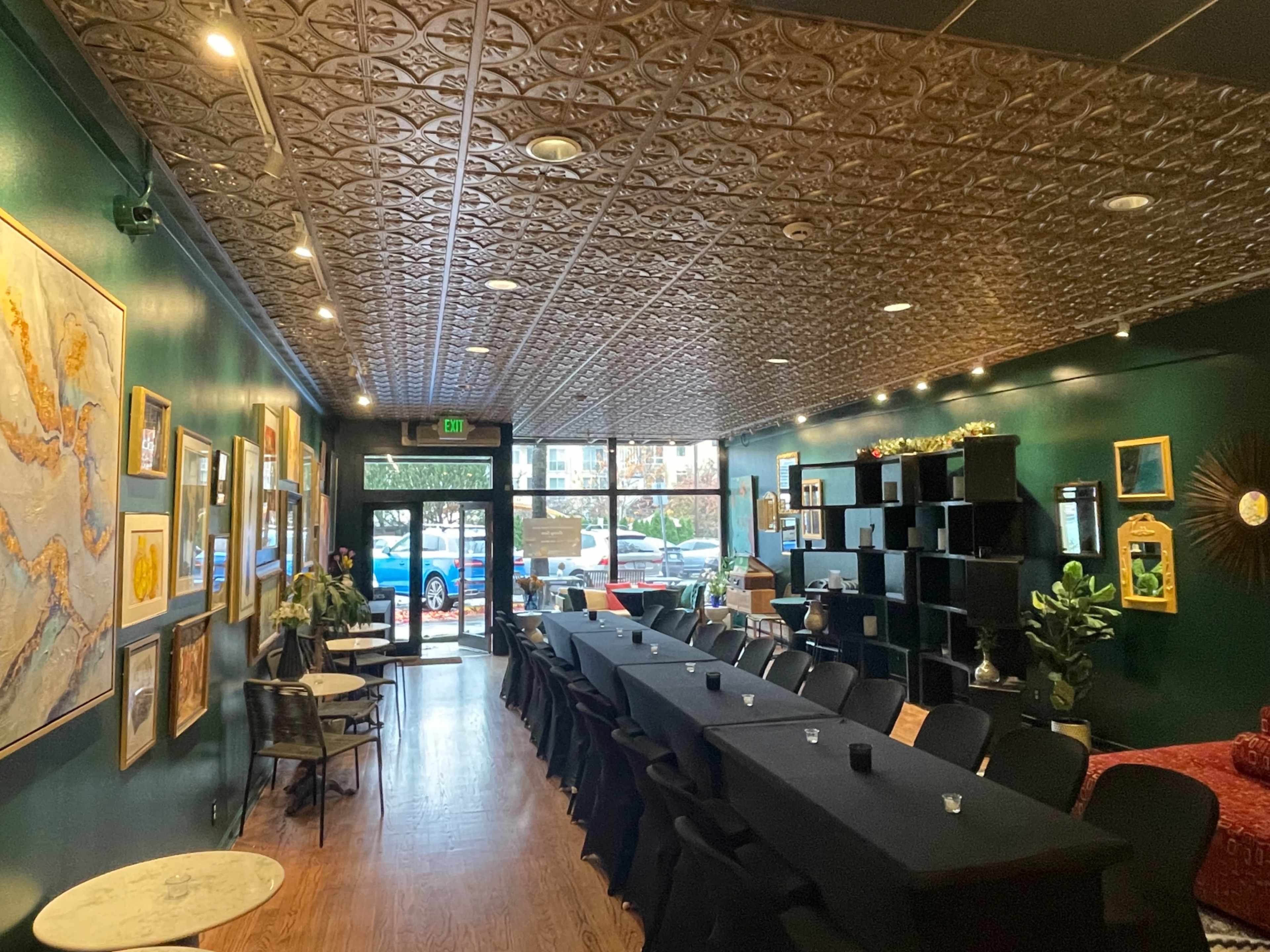 The image shows a restaurant interior featuring dark green walls, a patterned ceiling, and a long table with black cloth, surrounded by black chairs and decorative artwork on the walls.