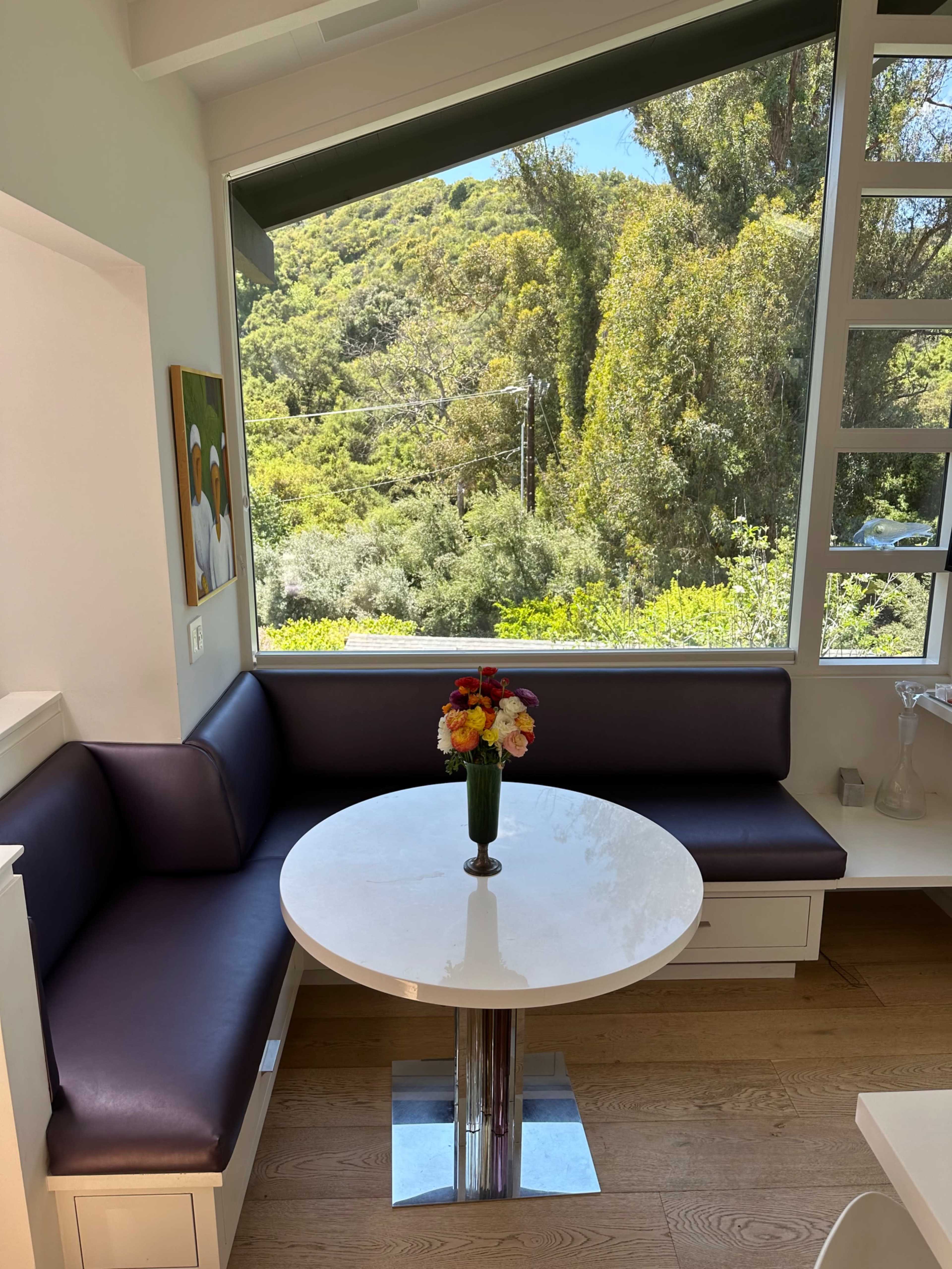 A round table with a vase of flowers sits in a corner nook beside a large window overlooking a green landscape.