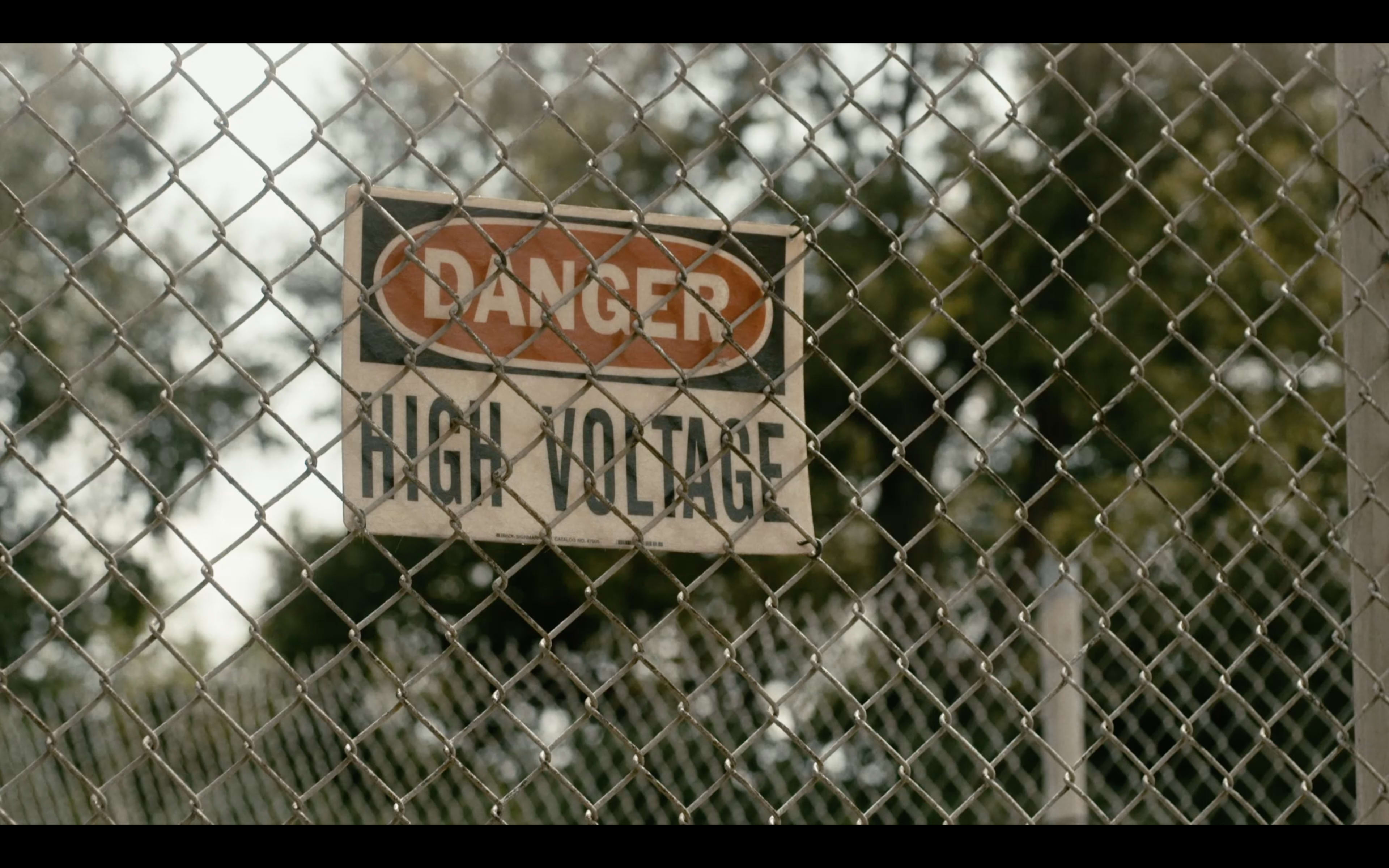 A warning sign indicating "DANGER HIGH VOLTAGE" is attached to a chain-link fence in a wooded area.